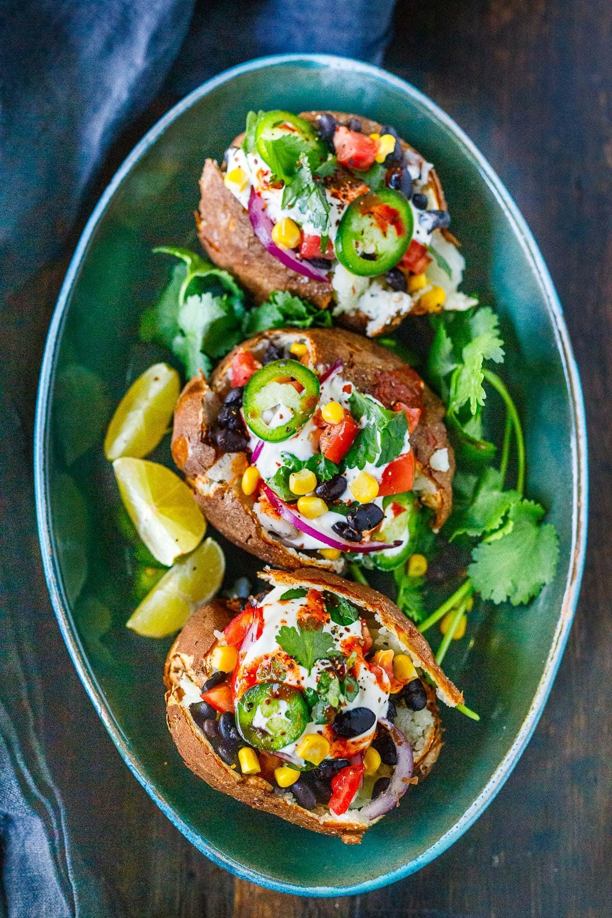 three Mexican stuffed baked potatoes on a plate with fresh cilantro and lime, topped with corn, black beans, red onion, diced tomatoes, sliced jalapeño, with sour cream.