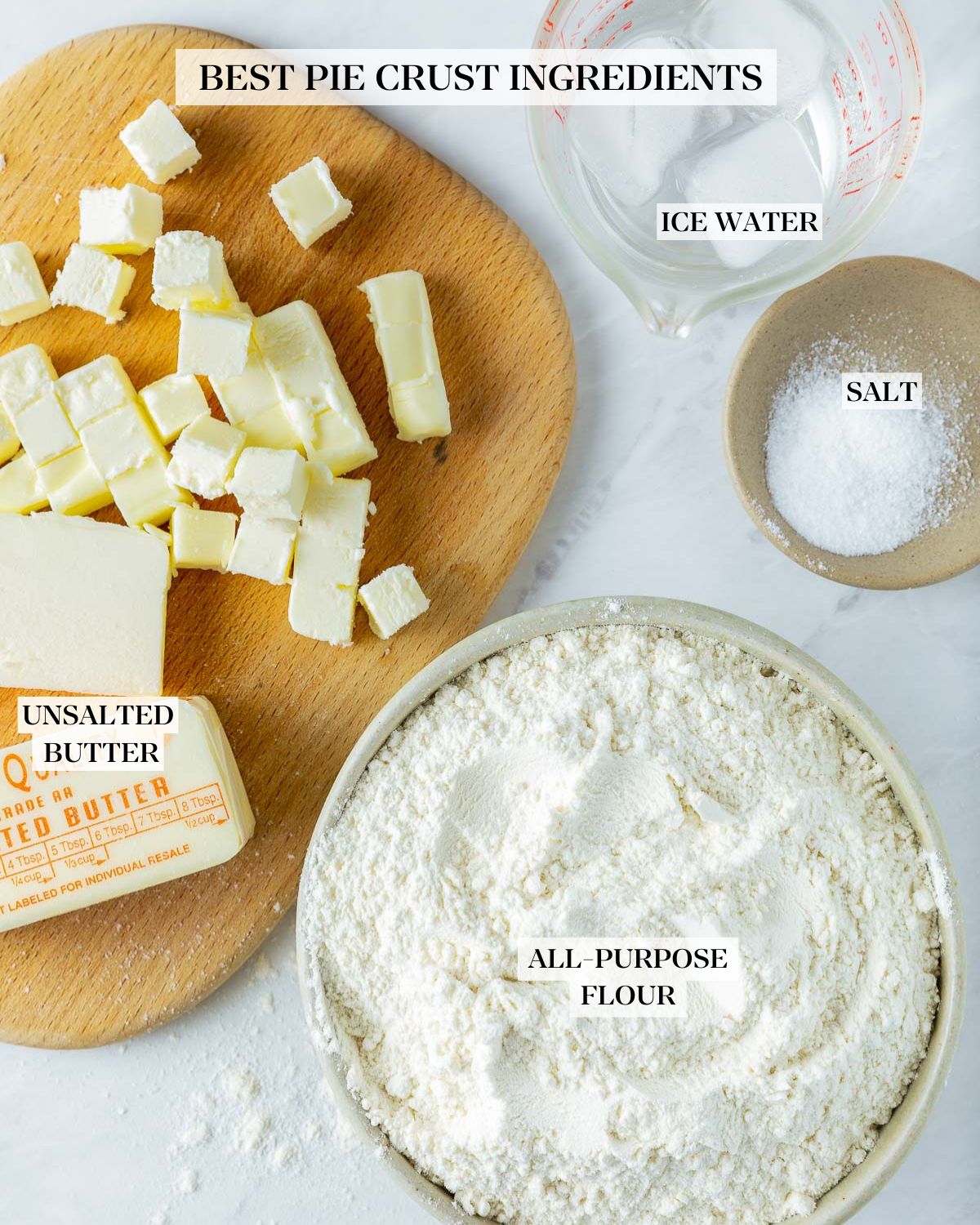 pie crust ingredients laid out - measuring cup with ice water, small bowl with salt, wood board with cubed butter, butter stick, and bowl of flour. 