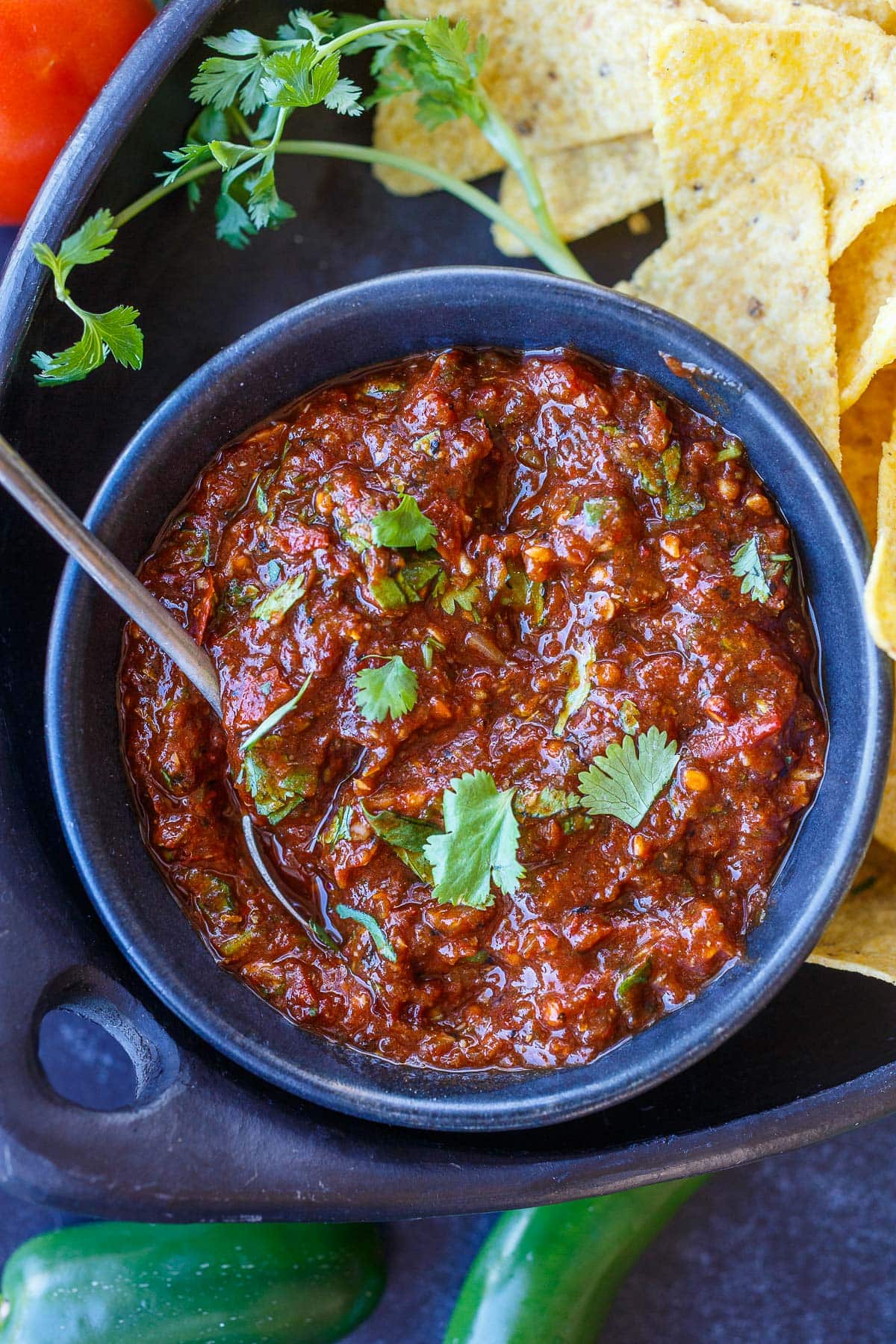 bowl of salsa roja topped with cilantro with spoon on plate. with tortilla chips.