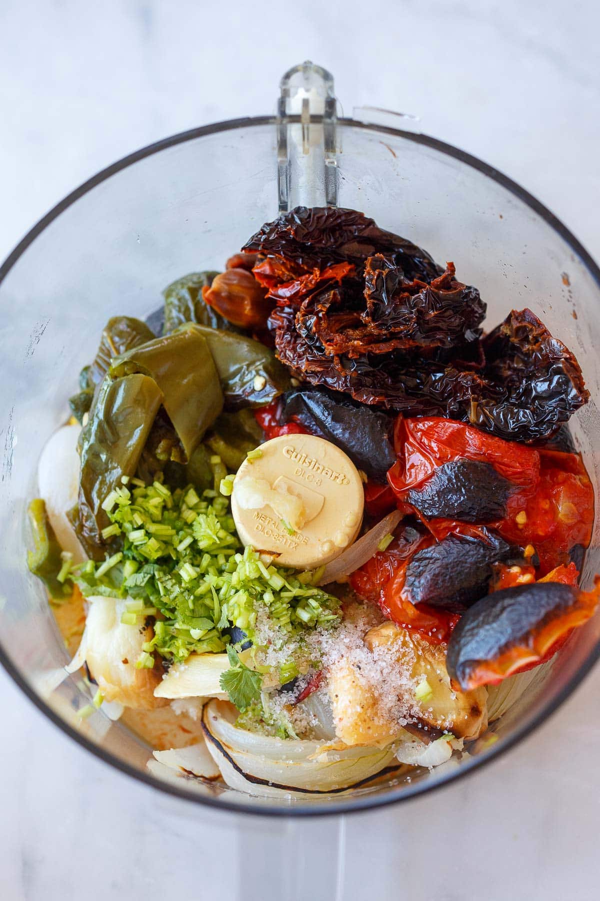 salsa roja ingredients in bowl of food processor - charred tomatoes, dried chilies, peppers, cilantro, salt, onion, and garlic.