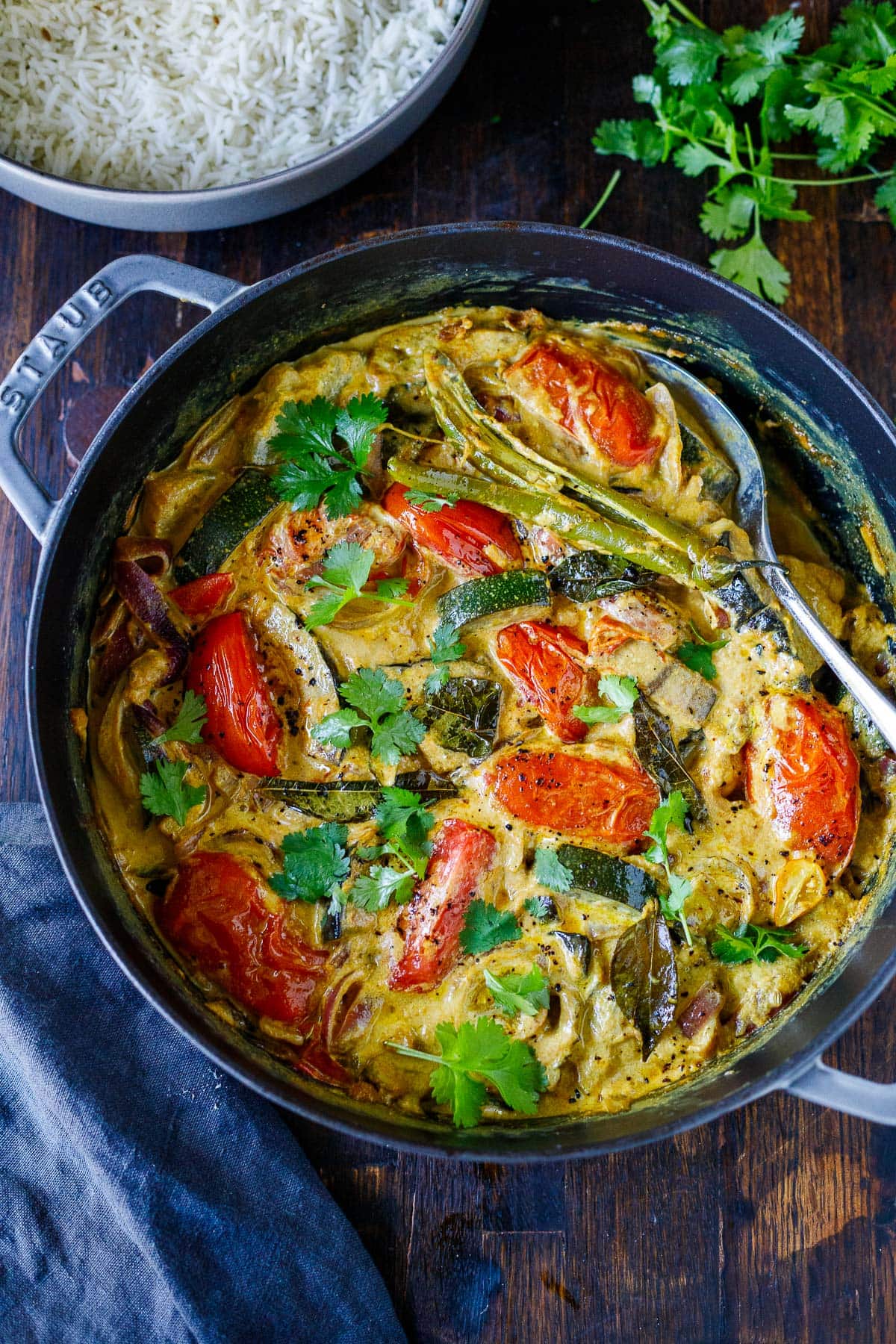 large pot of vegetable curry with serving spoon, with tomatoes, zucchini, eggplant, onions, chilies, curry leaves, and fresh cilantro. 
