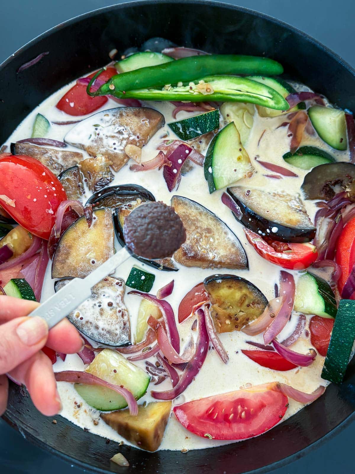 hand holding a teaspoon of tamarind paste above a large pot with vegetable curry in coconut milk. 