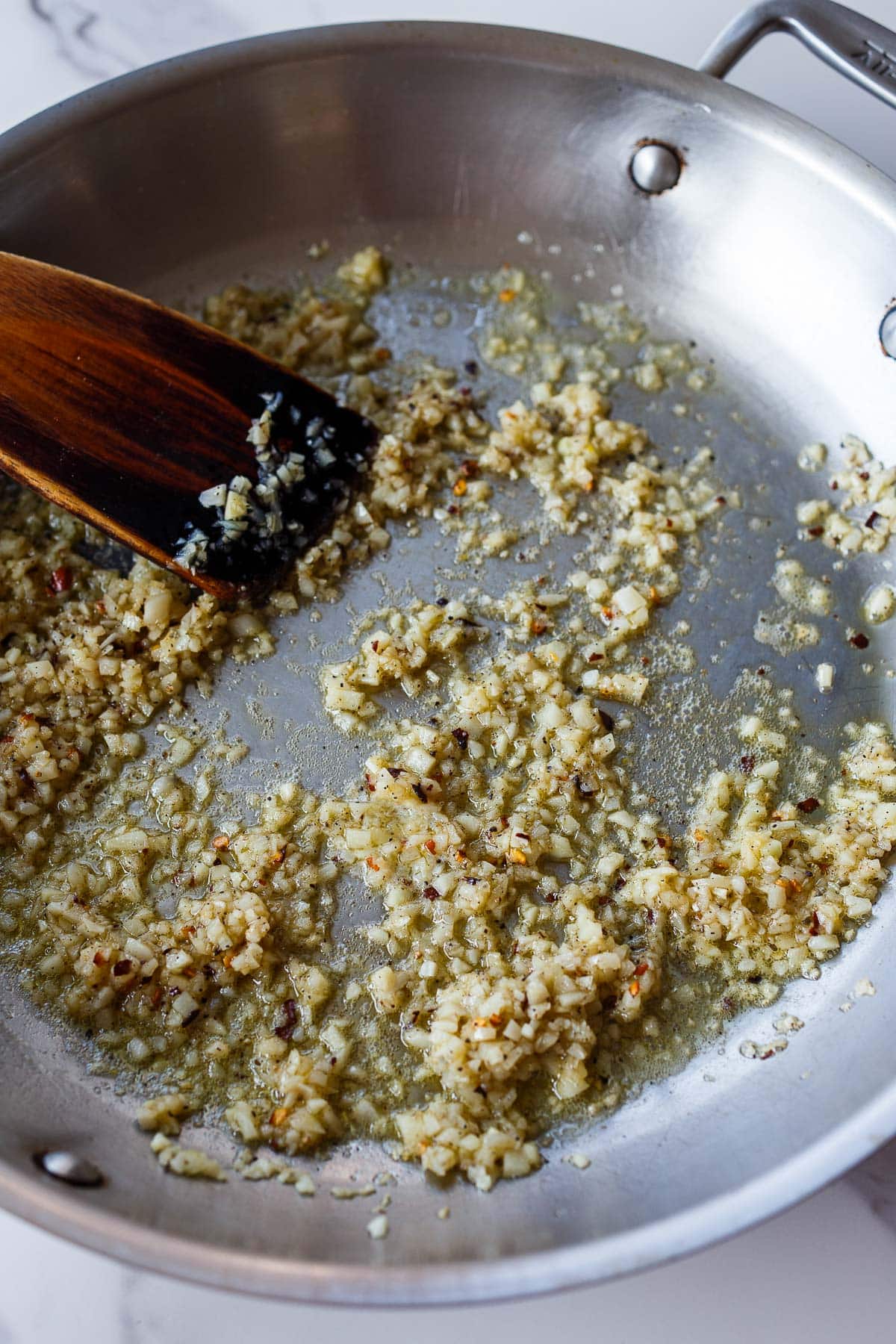 minced garlic and chili flakes sautéing in olive oil in a pan with a wood spatula. 