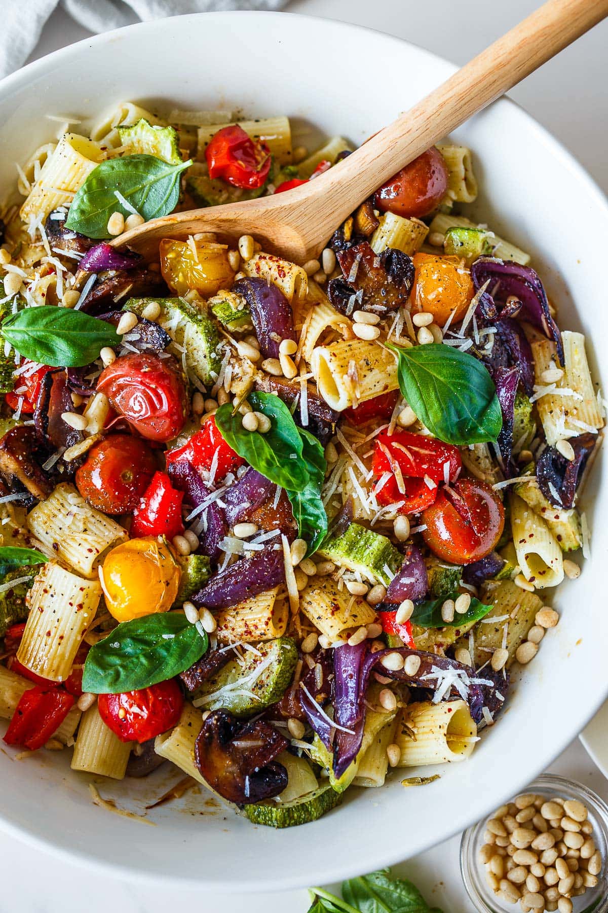 roasted vegetable pasta with cherry tomatoes, red onion, mushrooms, zucchini, and bell peppers, garnished with basil, parmesan, and pine nuts in large bowl. 