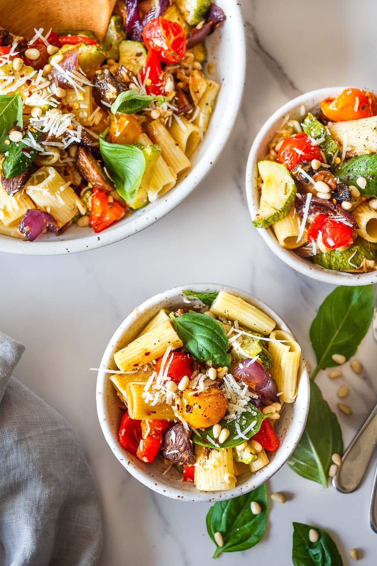 two small bowls of roasted vegetable pasta with parmesan, pine nuts, and basil next to large bowl of roasted vegetable pasta. 