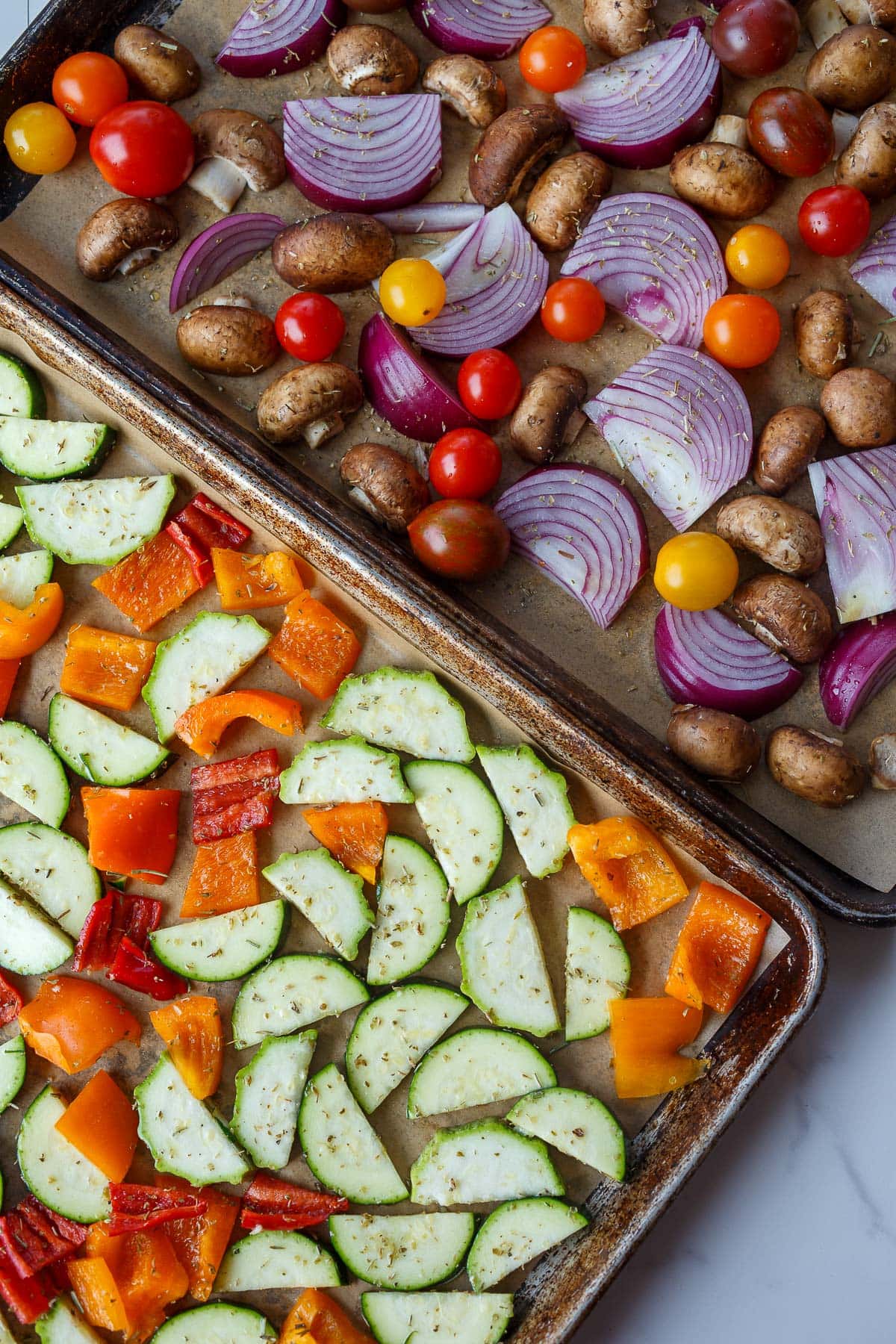 two baking sheets with cut veggies spread out - zucchini and bell peppers on one and red onion, tomatoes, and mushrooms on the other. 