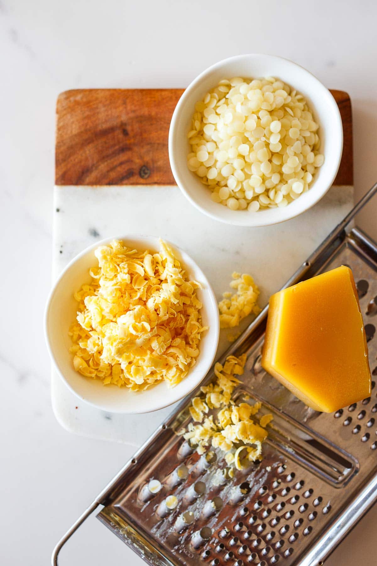 two bowls - one with grated beeswax, and the other with beeswax granules, next to grater with bar of beeswax. 