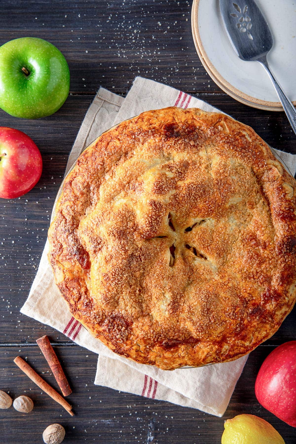 classic apple pie with golden topping with coarse sugar with five slits in shape of star next to apples and cinnamon sticks.