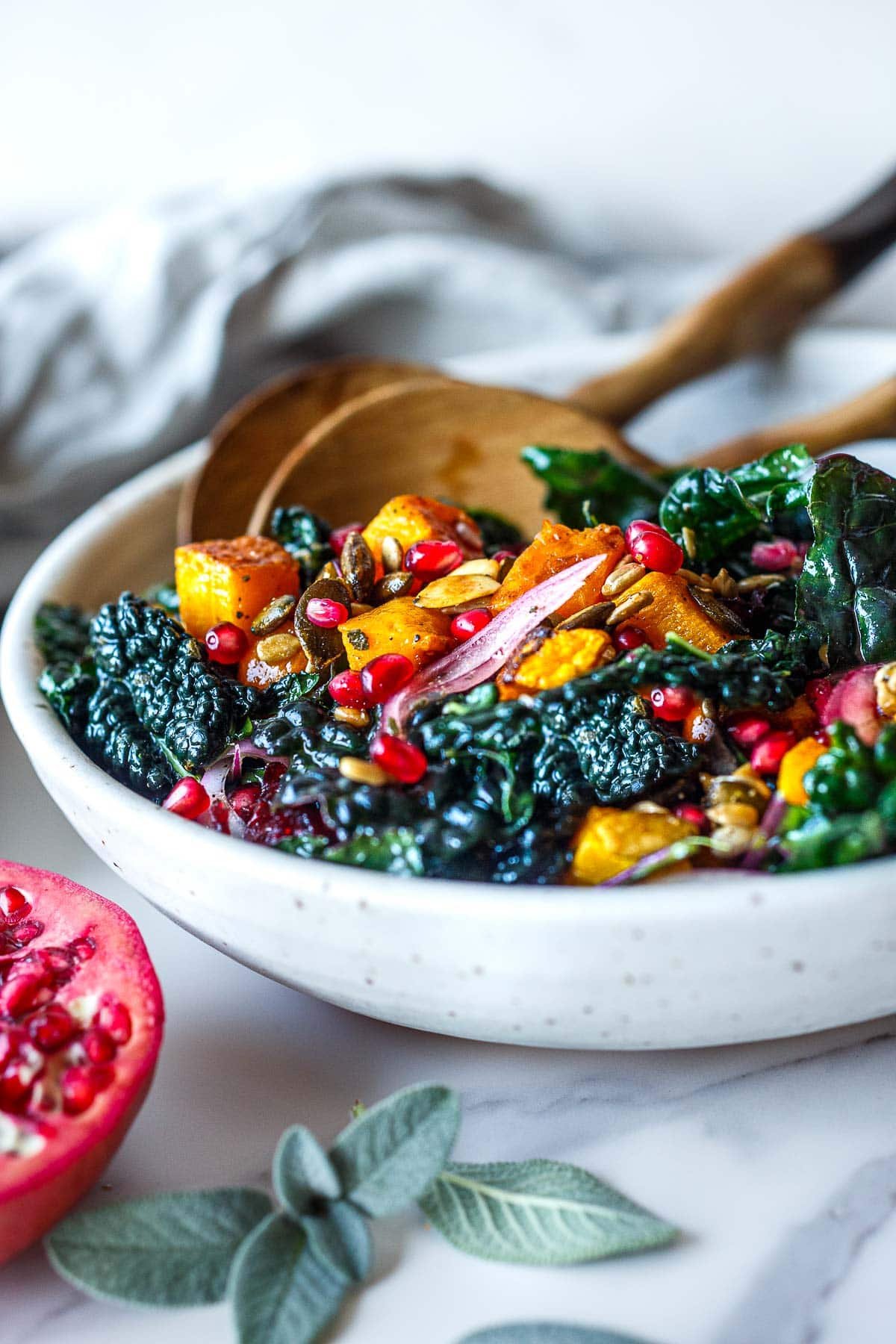 roasted butternut squash kale salad with sliced red onion, pomegranate arils, toasted seeds, and wood salad utensils.