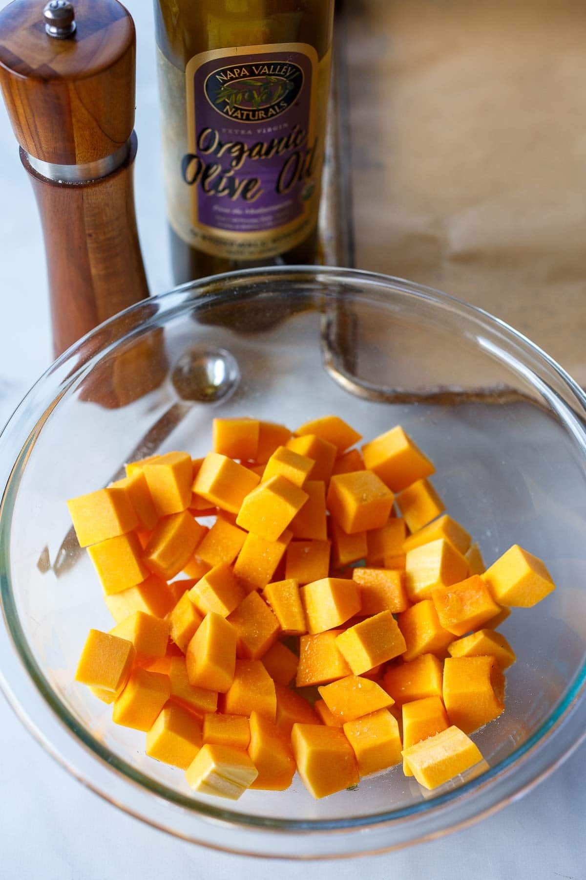 glass mixing bowl with cubed butternut squash with salt shaker and bottle of olive oil behind.