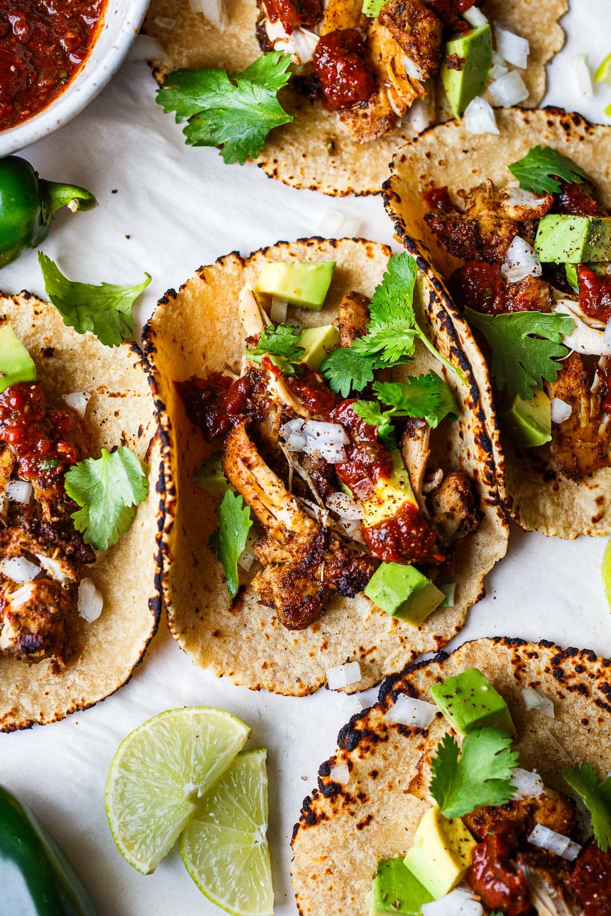 five chicken tacos topped with salsa roja, cilantro, and avocado, next to lime halves, jalapeños, bowl of salsa, and avocado half.