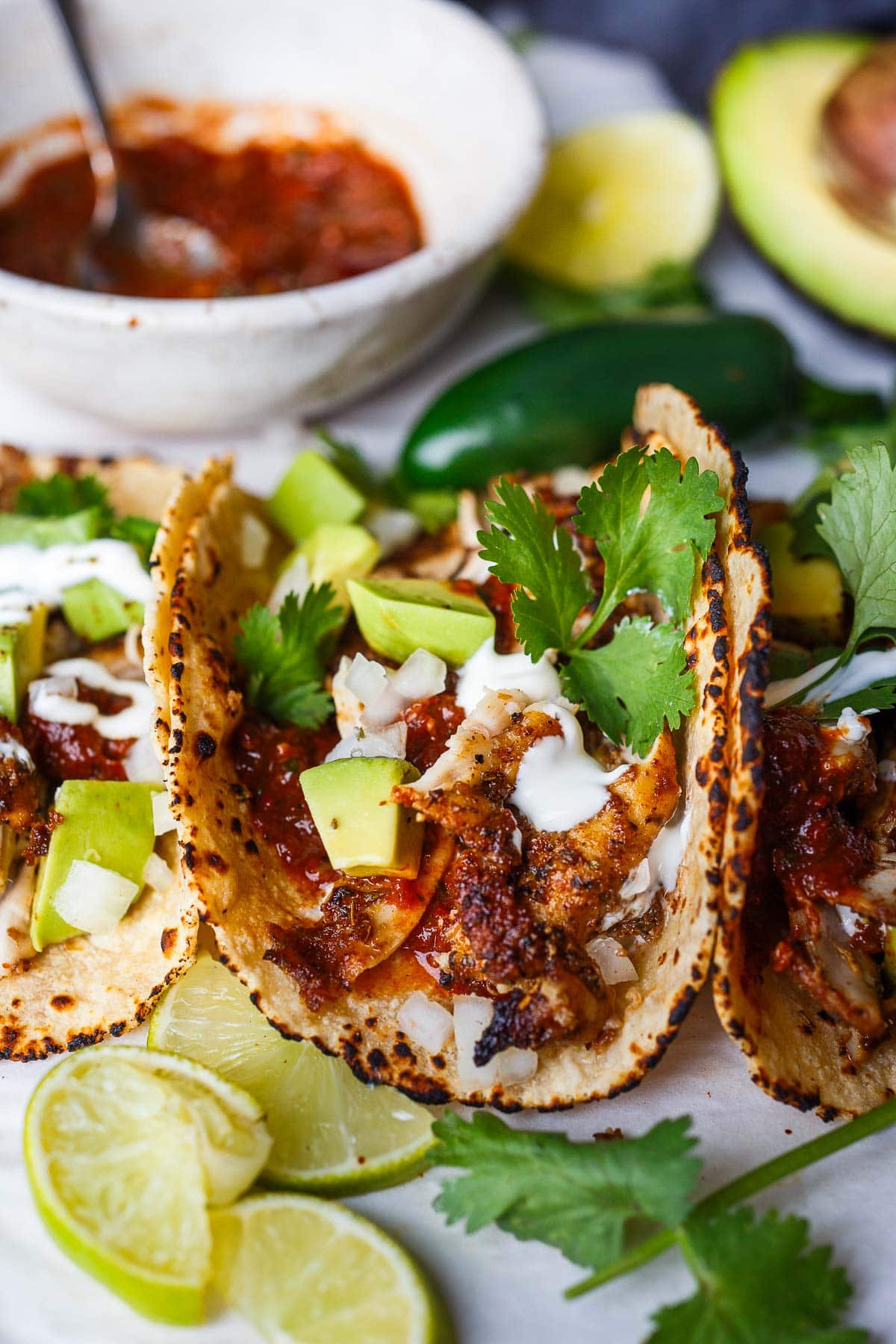 chicken taco recipe topped with salsa roja, cilantro, and avocado, next to lime halves, jalapeños, bowl of salsa, and avocado half.