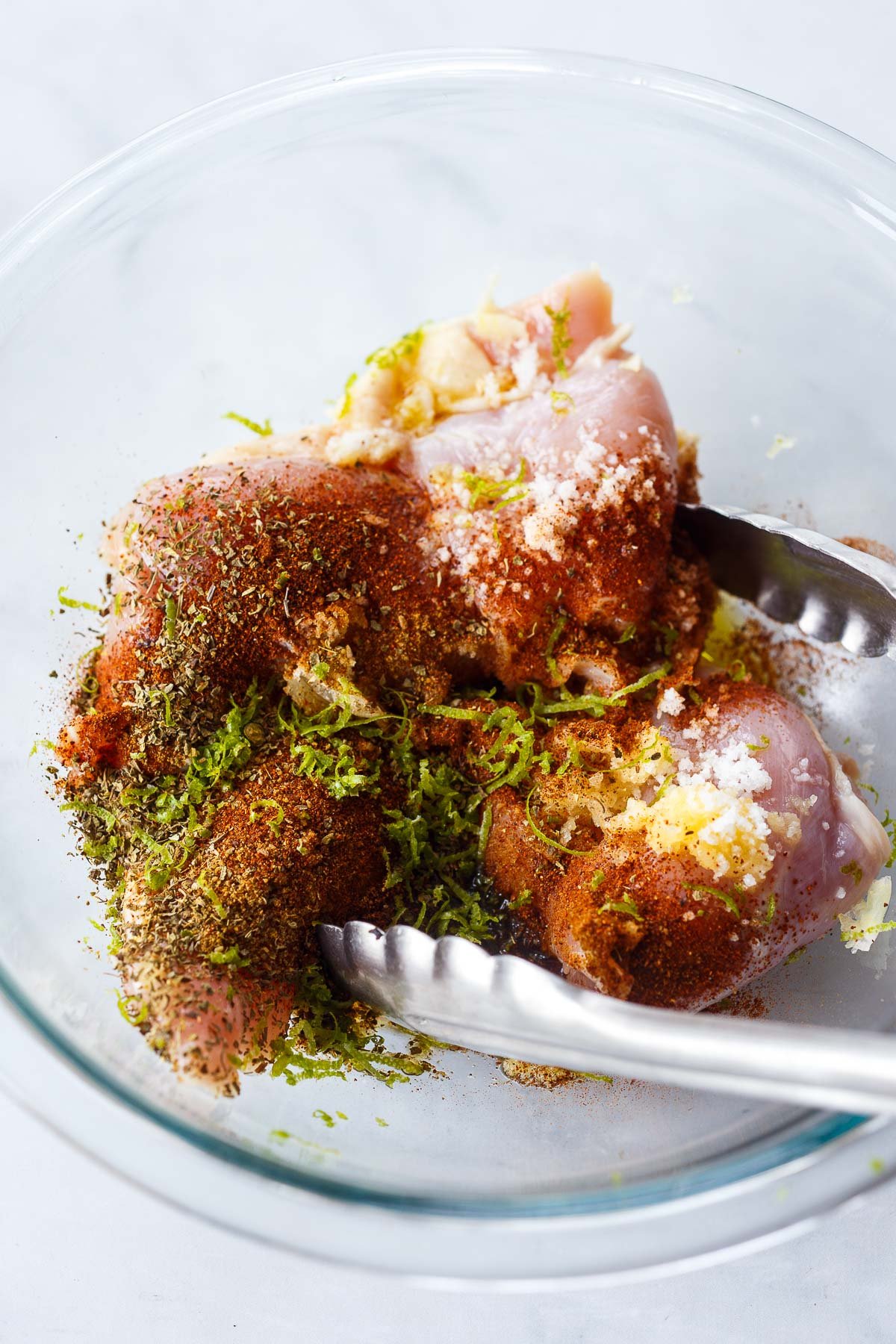 glass mixing bowl with chicken thighs in spices, lime zest, and garlic marinade, with tongs to toss.