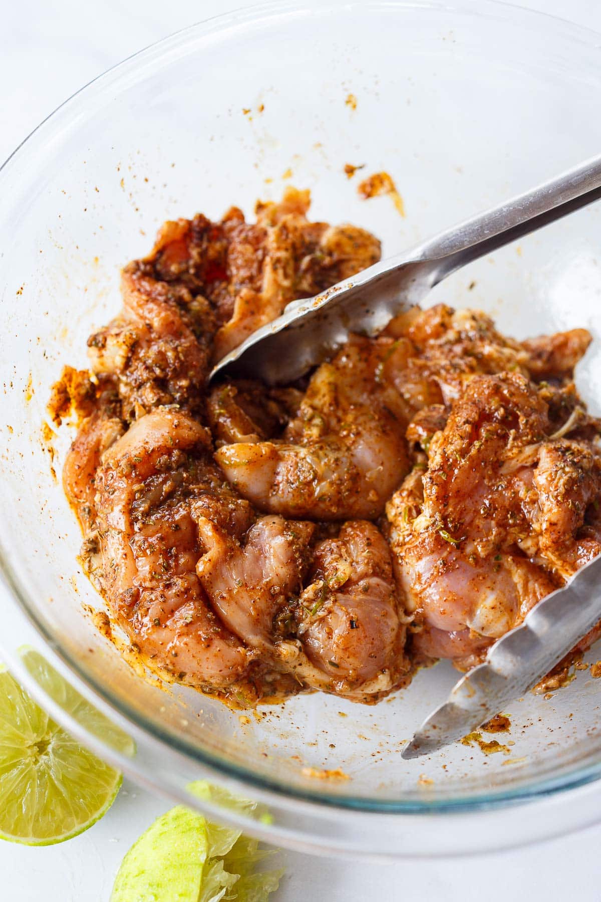 glass mixing bowl with chicken thighs tossed in marinade with tongs.