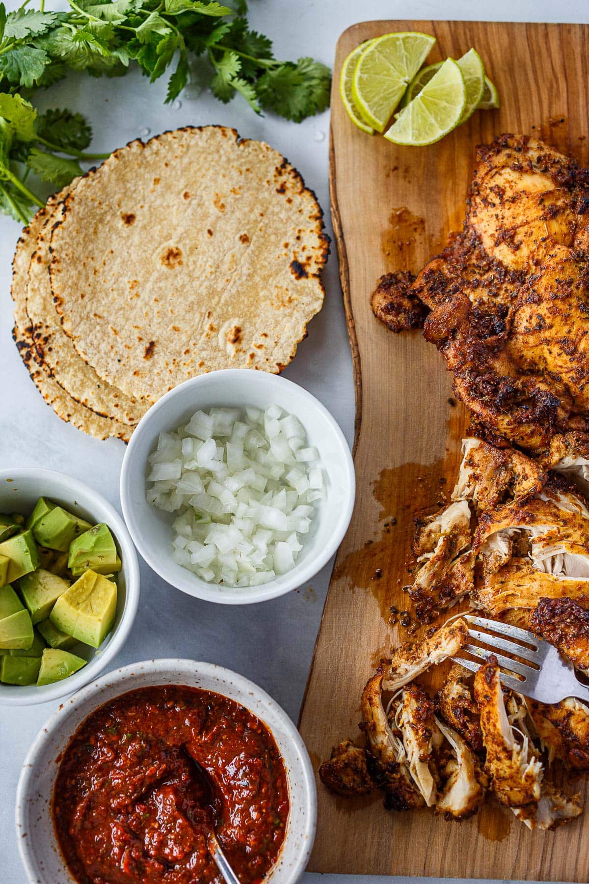 components for chicken tacos - marinated baked chicken on wood board, lime halves, corn tortillas, diced white onion, diced avocado, salsa roja. 