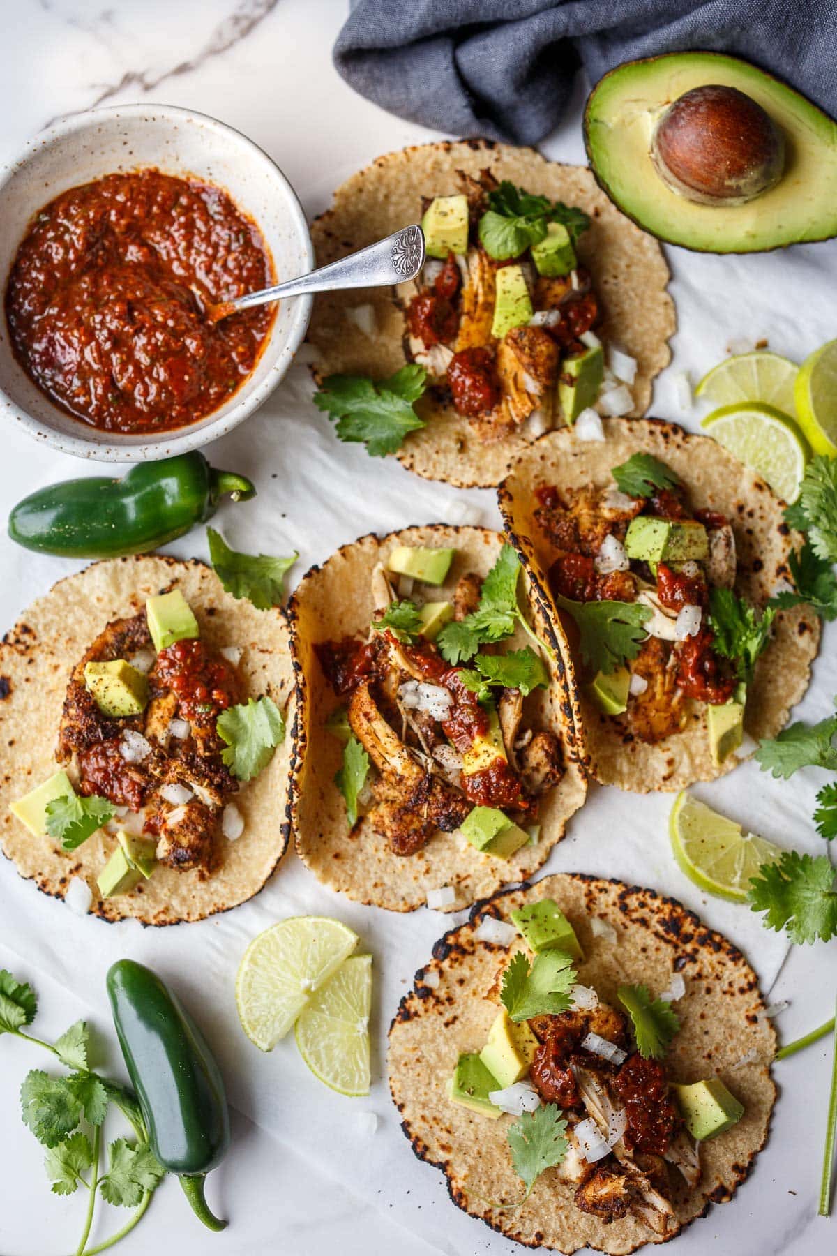 five chicken tacos topped with salsa roja, cilantro, and avocado, next to lime halves, jalapeños, bowl of salsa, and avocado half.