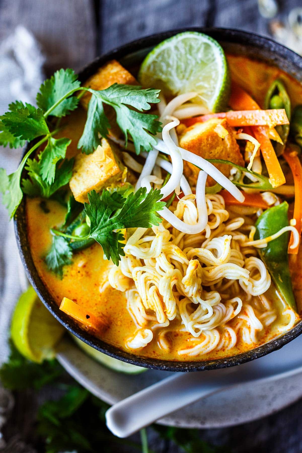 bowl of curry ramen with cilantro, seared tofu cubes, carrots, snap peas, and bean sprouts with a lime wedge.