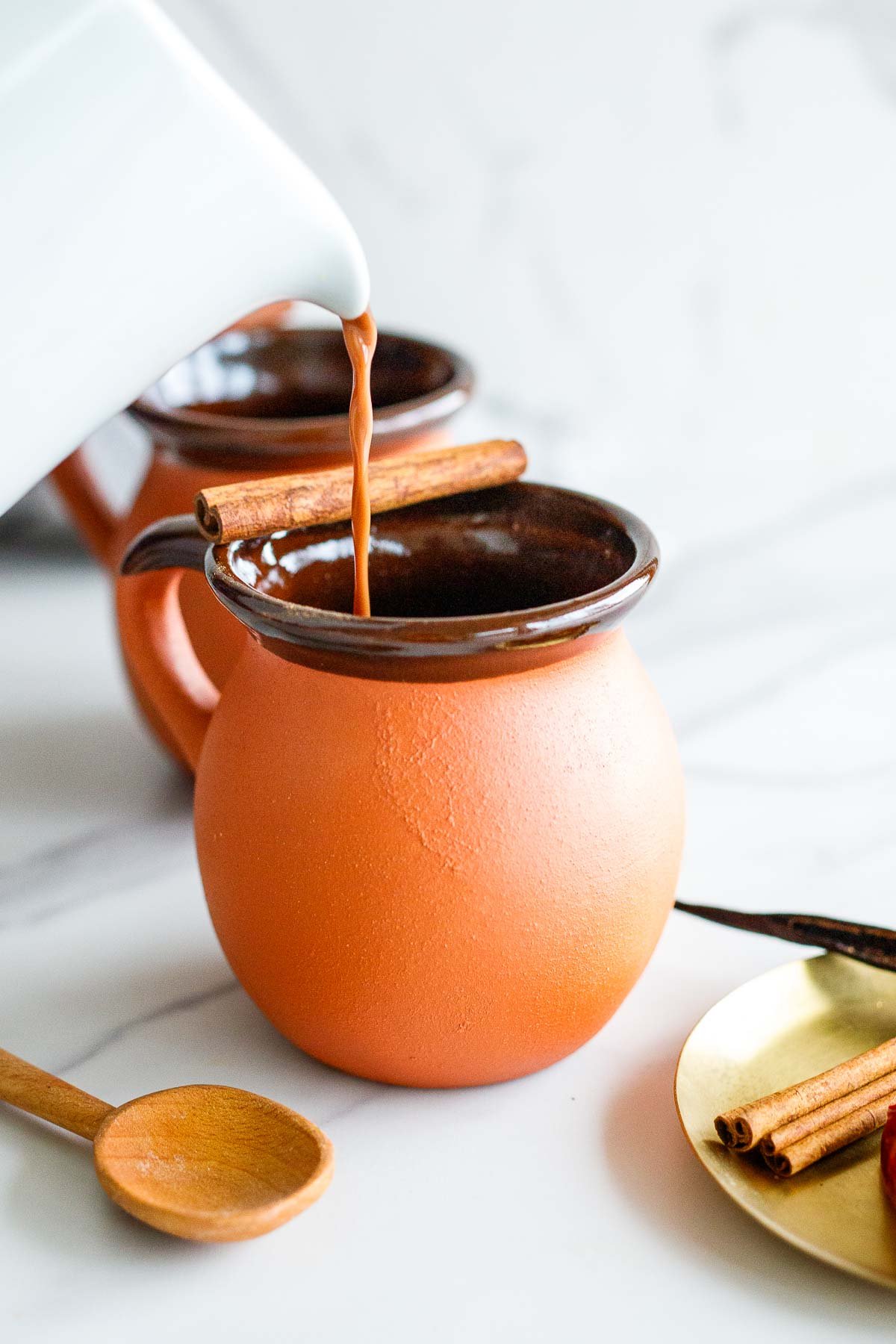 pouring Mexican hot chocolate into a ceramic mug with a cinnamon stick resting on the edge.