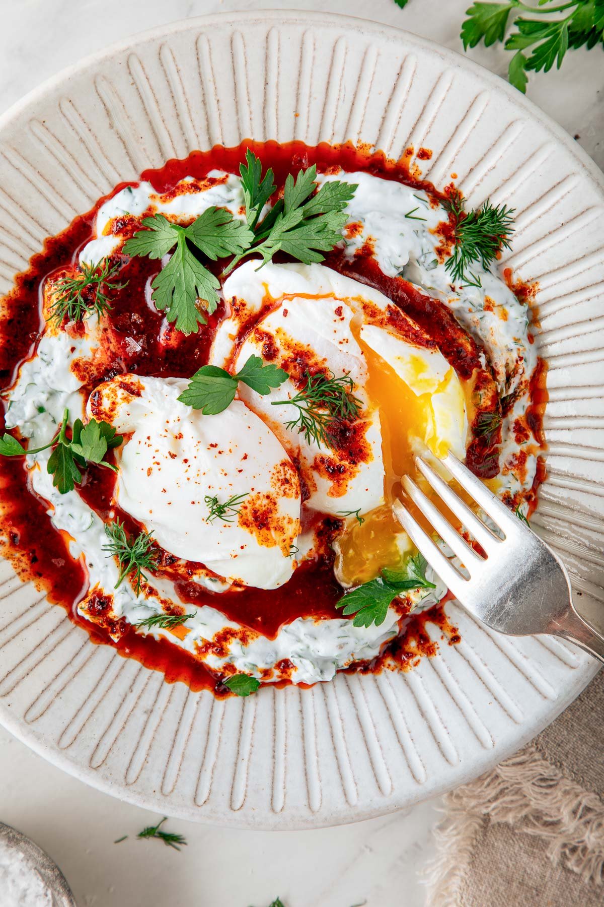 Turkish eggs on plate with fork breaking open poached eggs, garnished with fresh herbs and drizzle of Aleppo butter. 