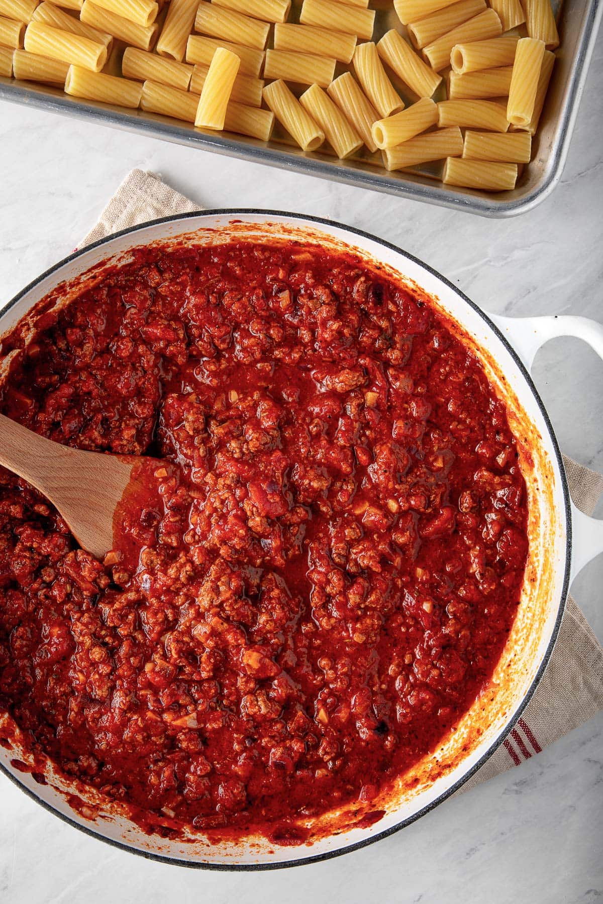 dutch oven with tomato meat sauce with wooden spatula next to baking sheet with cooked rigatoni spread out.