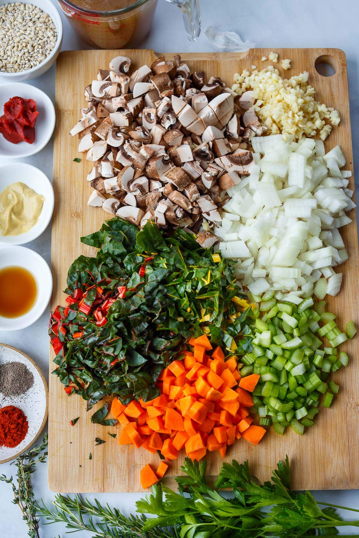 mise en place - wood board with minced carrots, celery, onion, garlic, mushrooms, and chard next to small bowls of barley, tomato paste, Dijon, vinegar, and spices.
