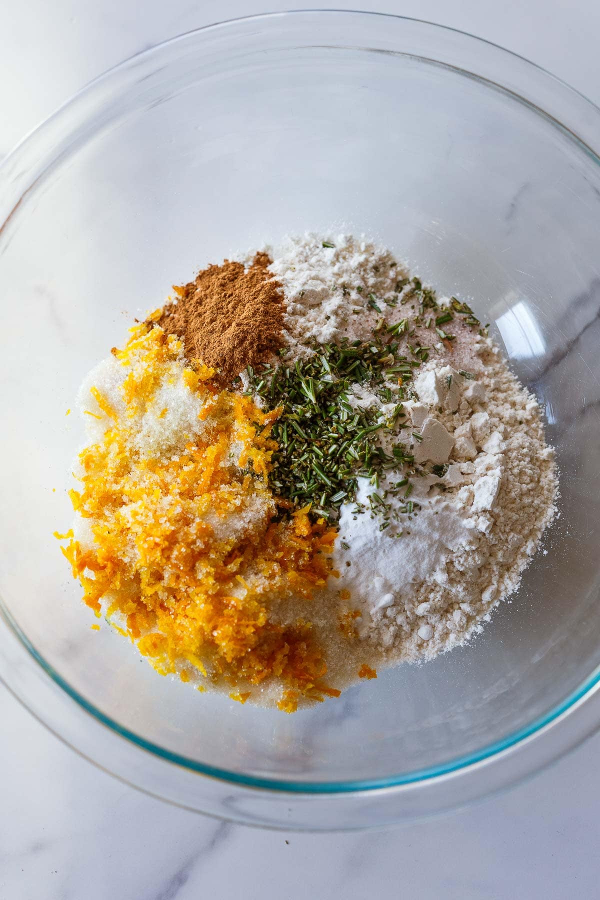 glass mixing bowl with dry ingredients for cranberry orange muffins - flour, salt, sugar, orange zest, rosemary.