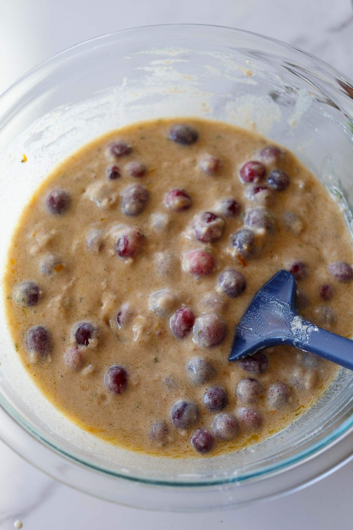 fresh cranberries in cranberry orange muffin batter in glass mixing bowl with silicone spatula. 