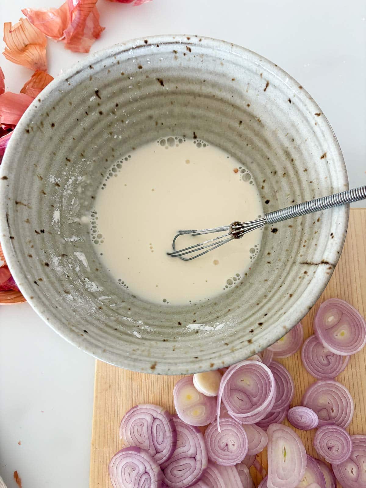 ceramic bowl with batter for frying shallots next to wood board of sliced shallots. 