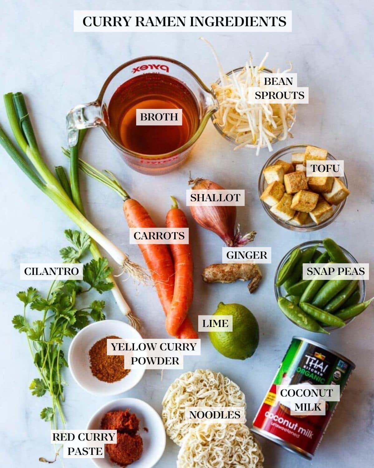 ingredients for curry ramen - measuring cup of broth, bowls of bean sprouts, cubed seared tofu, snap peas, coconut milk can, instant ramen noodles, lime, ginger, shallot, carrots, green onions, cilantro, and two small bowls of curry powder and curry paste.