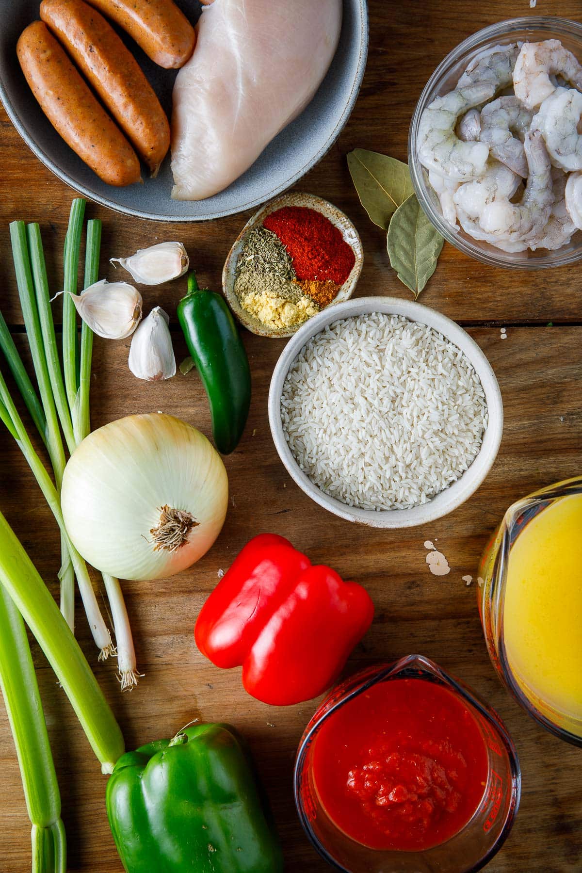 ingredients for jambalaya - plate of chicken andouille sausage, chicken, small bowls of shrimp, spices, rice, and bay leaves, garlic, jalapeno, green onions, onion, bell peppers, celery, and measuring cups with broth and crushed tomatoes.