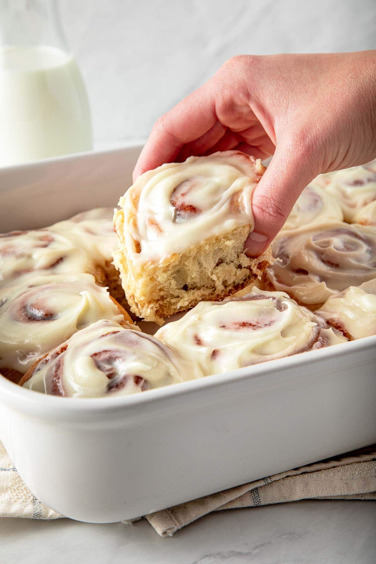 hand lifting a sourdough cinnamon roll out of a baking dish, with cream cheese frosting.