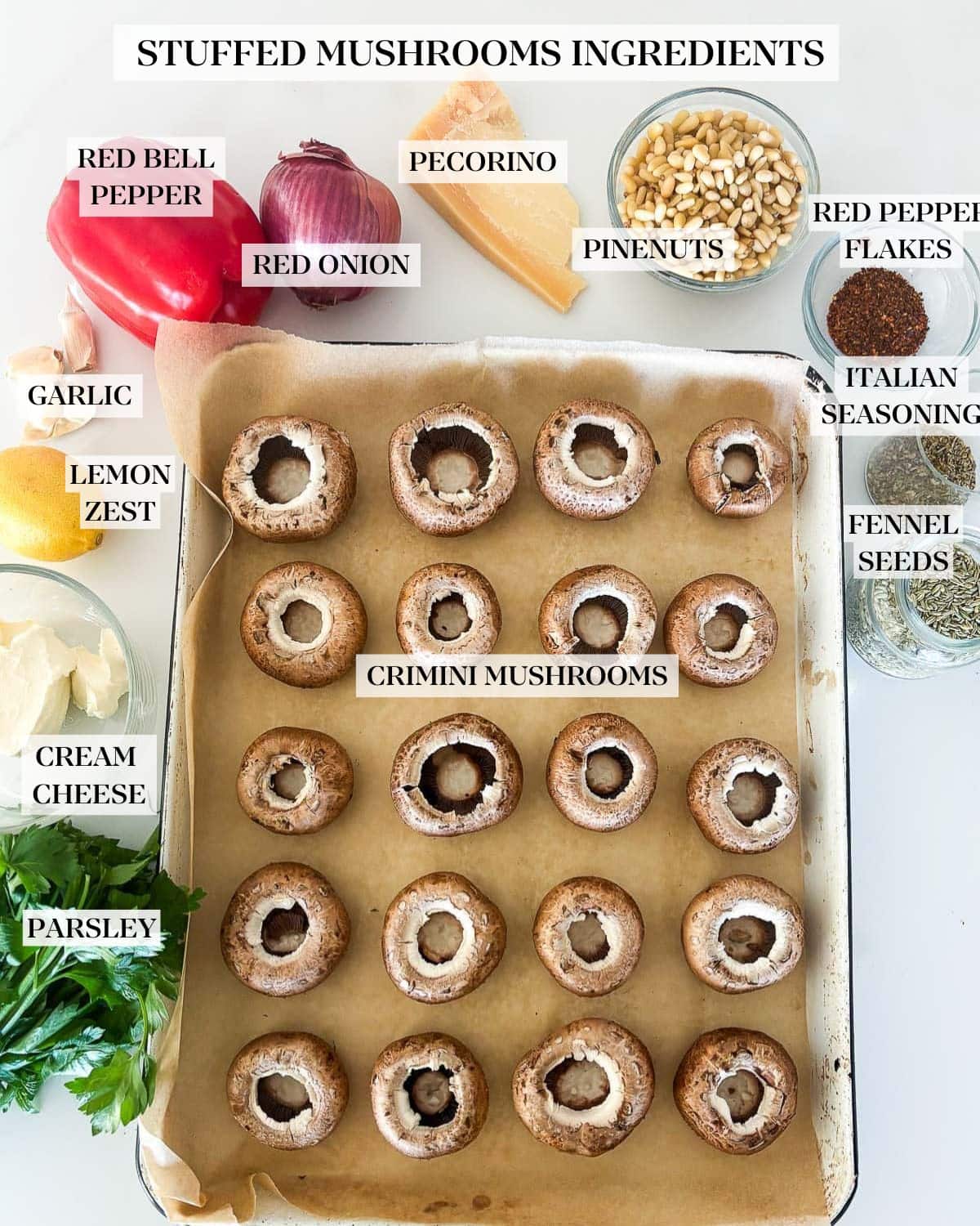 ingredients for stuffed mushrooms - parchment-lined sheet pan with mushroom caps, glass bowl of cream cheese, lemon, garlic cloves, red bell pepper, red onion, pecorino, pine nuts, red pepper flakes, dried Italian seasoning, fennel seeds, and fresh parsley. 