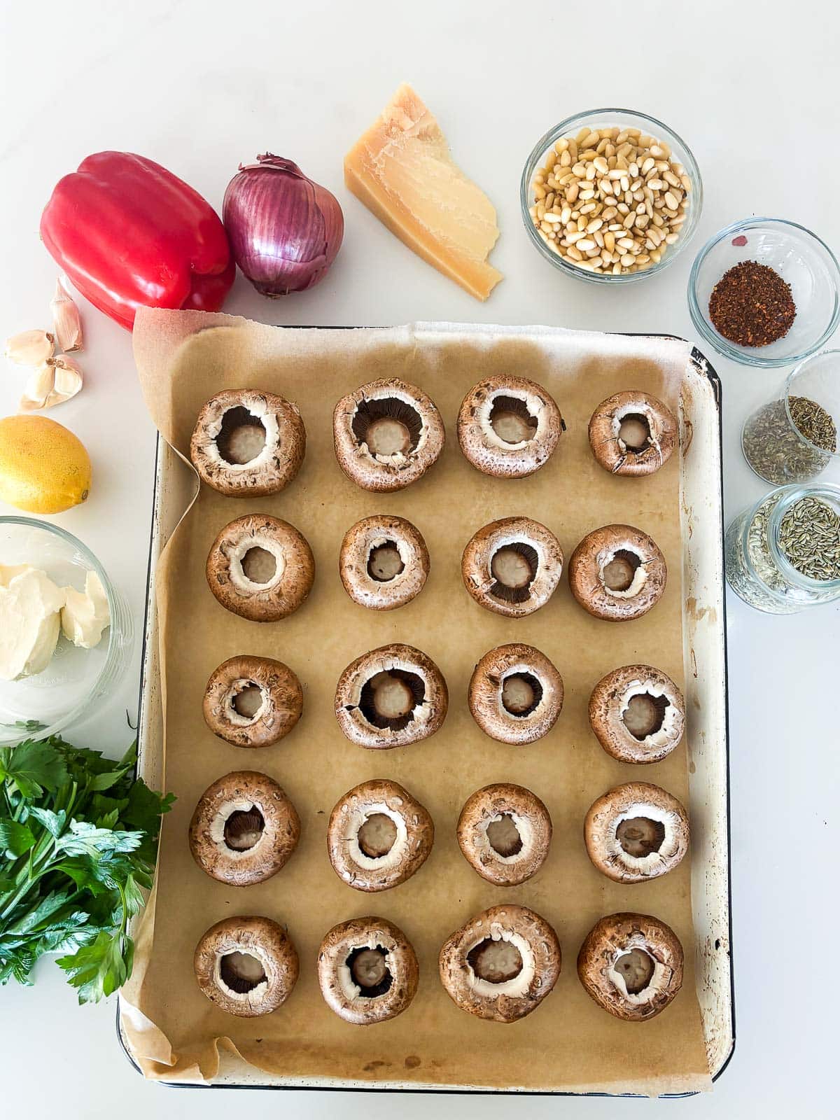 ingredients for stuffed mushrooms - parchment-lined sheet pan with mushroom caps, glass bowl of cream cheese, lemon, garlic cloves, red bell pepper, red onion, pecorino, pine nuts, red pepper flakes, dried Italian seasoning, fennel seeds, and fresh parsley.