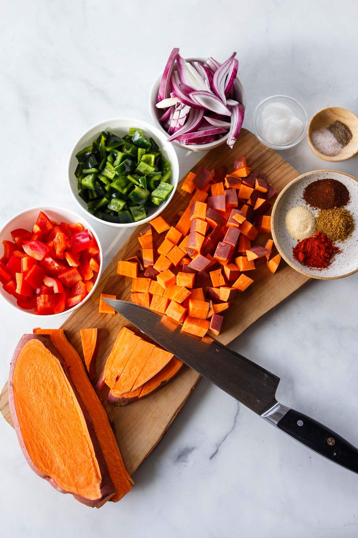 knife chopping sweet potato into 1/2-inch cubes on wood board, next to chopped red bell pepper, poblano, and onion in bowls, next to small dishes of oil, salt, pepper, and spices.