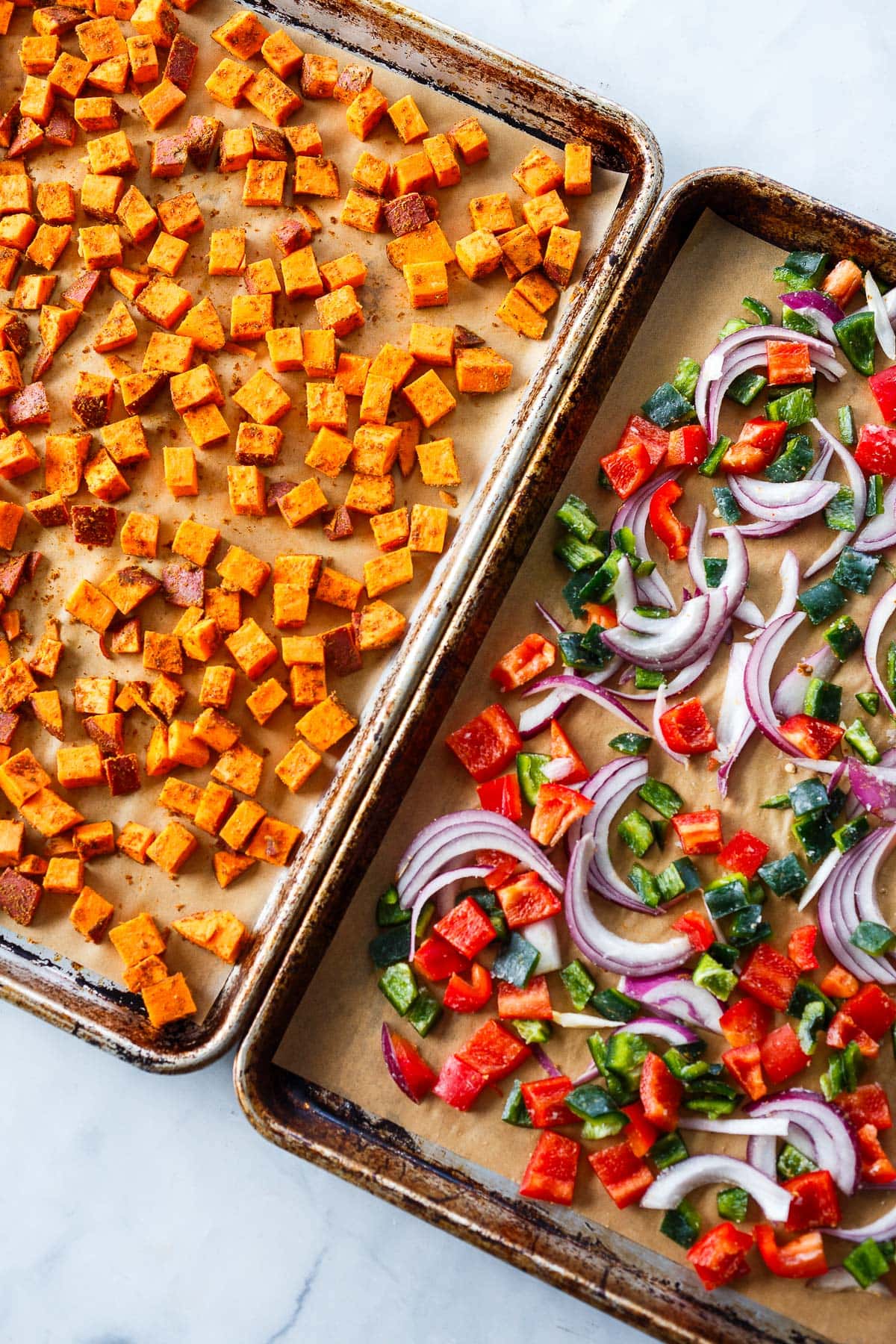 two baking sheets with parchment paper, one with cubed sweet potatoes, and one with red onion slices and diced peppers.