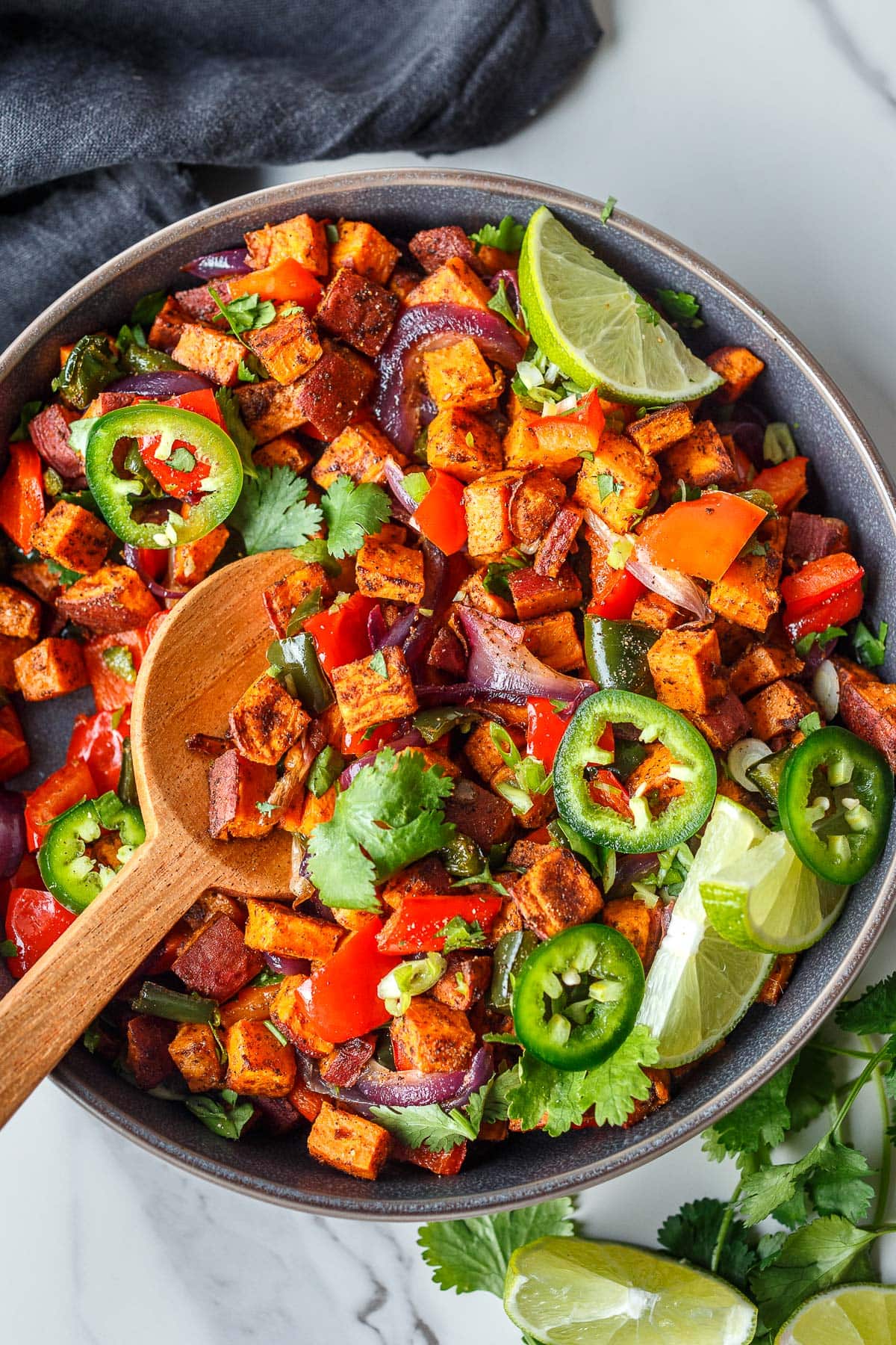 bowl of sweet potato hash with onions, peppers, sliced jalapeno, lime half moons, and a wood spoon.