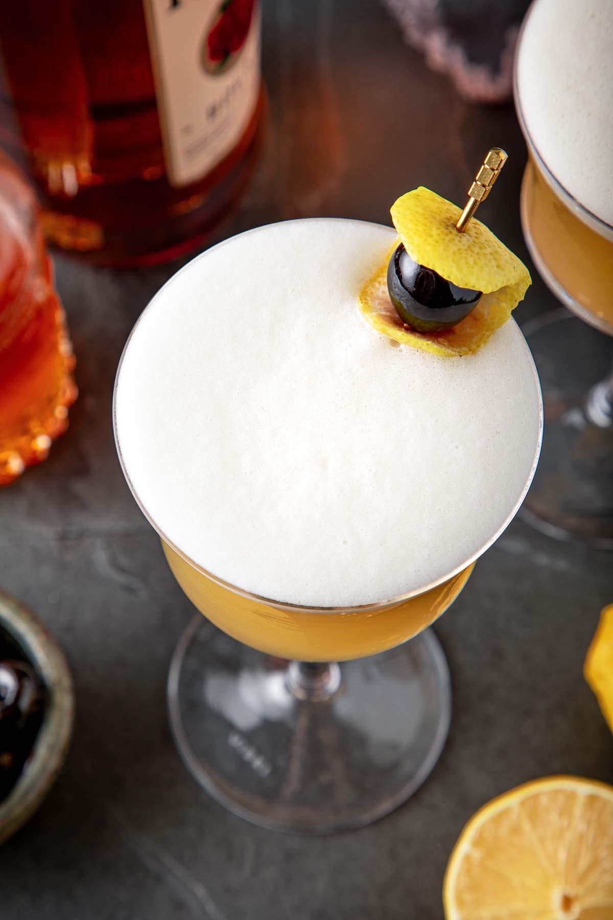 classic whiskey sour in coupe glass with frothy top with brass toothpick with lemon rind and Luxardo cherry. 