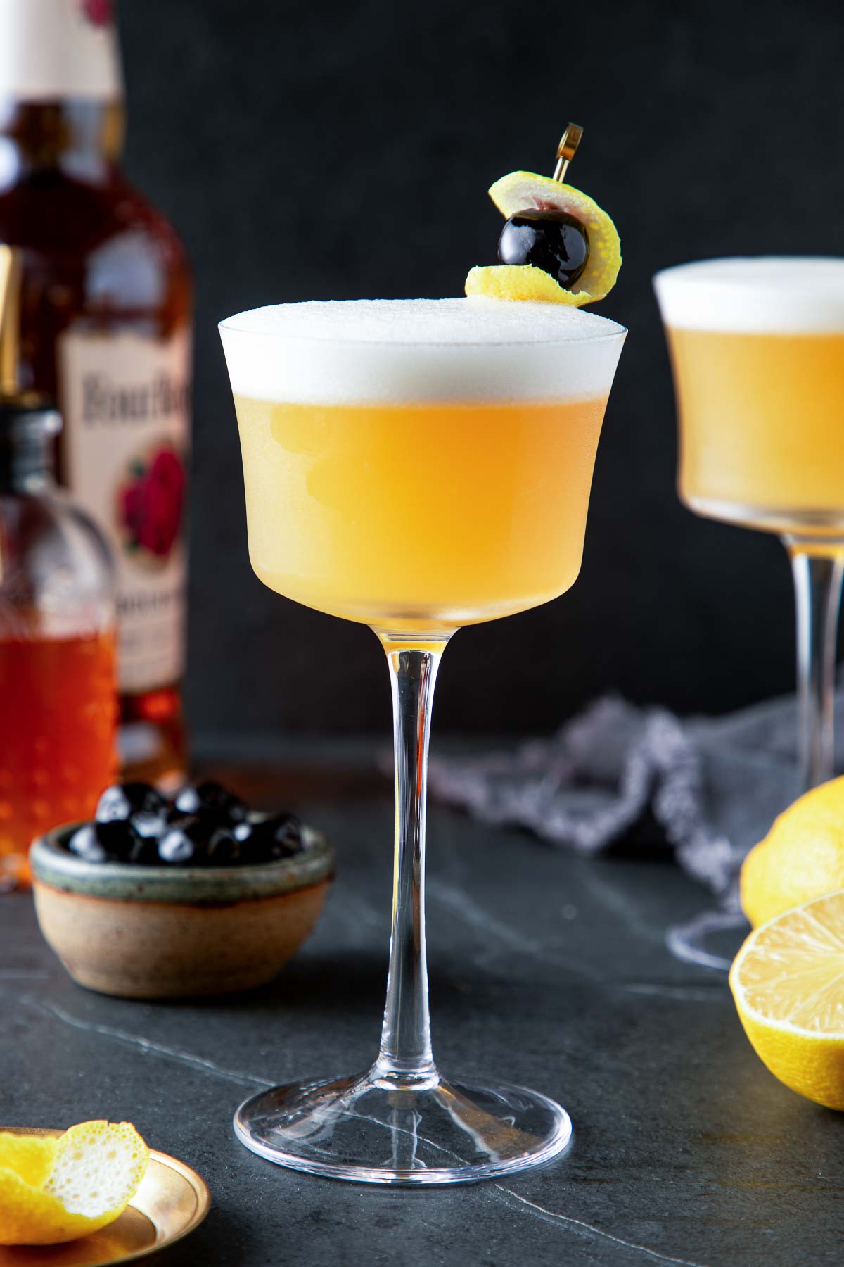 classic whiskey sour in a coupe glass with lemon rind and Luxardo cherry on brass toothpick. 