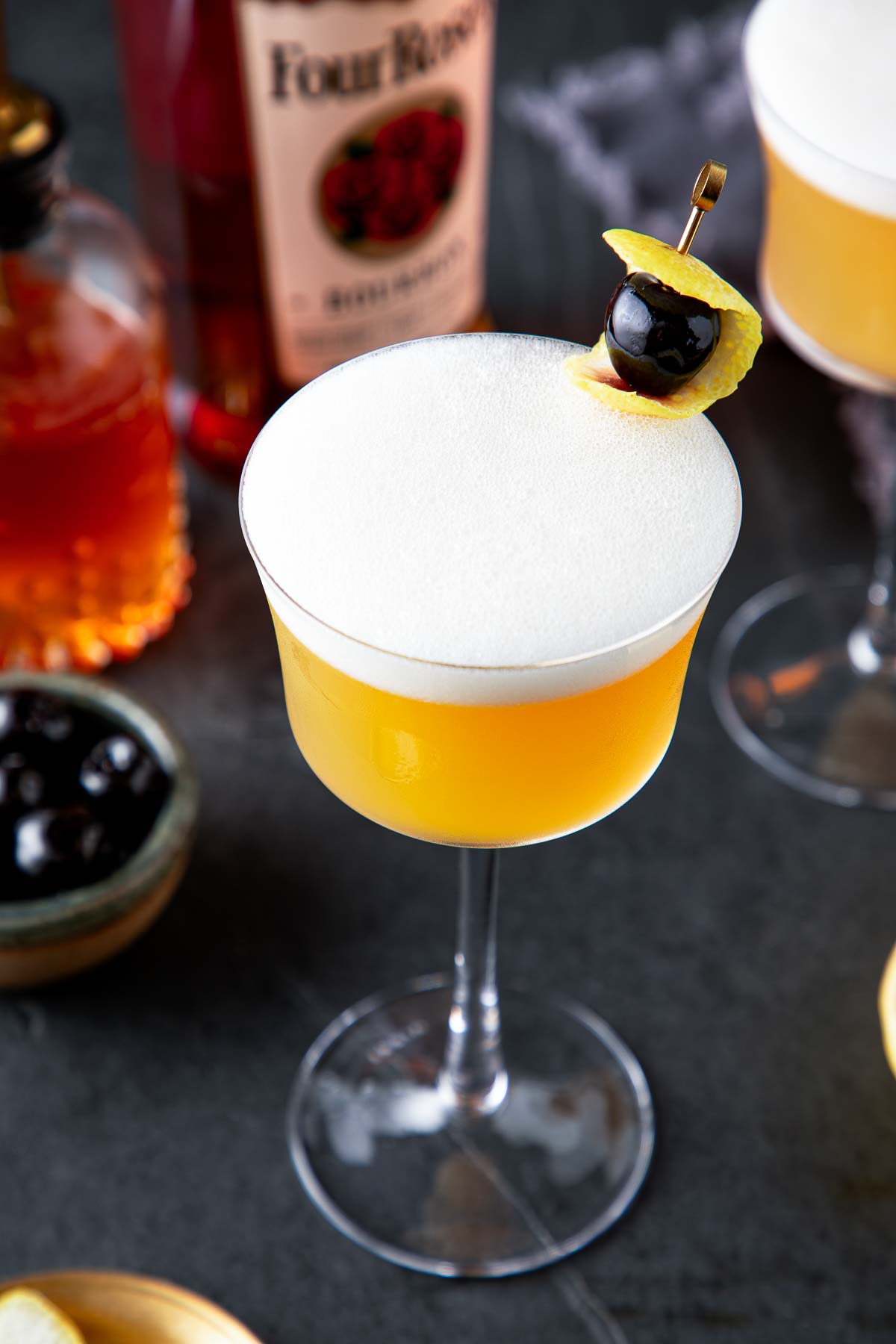 classic whiskey sour in coupe glass with frothy egg white top and lemon rind and Luxardo cherry for garnish on brass toothpick. 