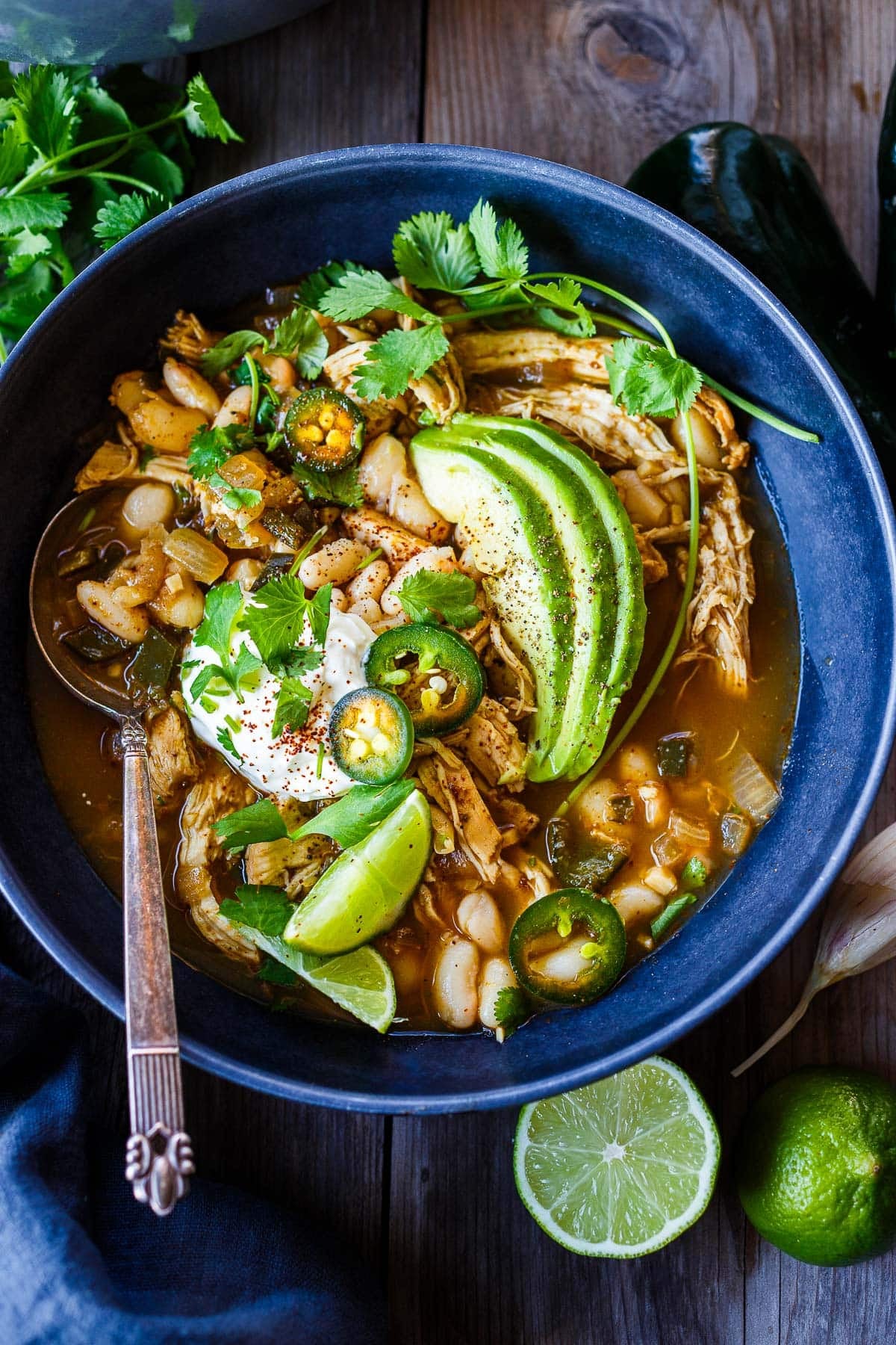 bowl of white bean chicken chili with sliced avocado, jalapeños, sour cream, lime wedges, and fresh cilantro, with spoon.