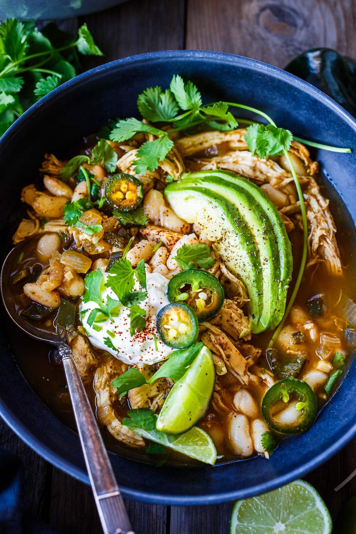 White chicken chili in a serving bowl with avocado, sour cream lime wedges and cilantro.