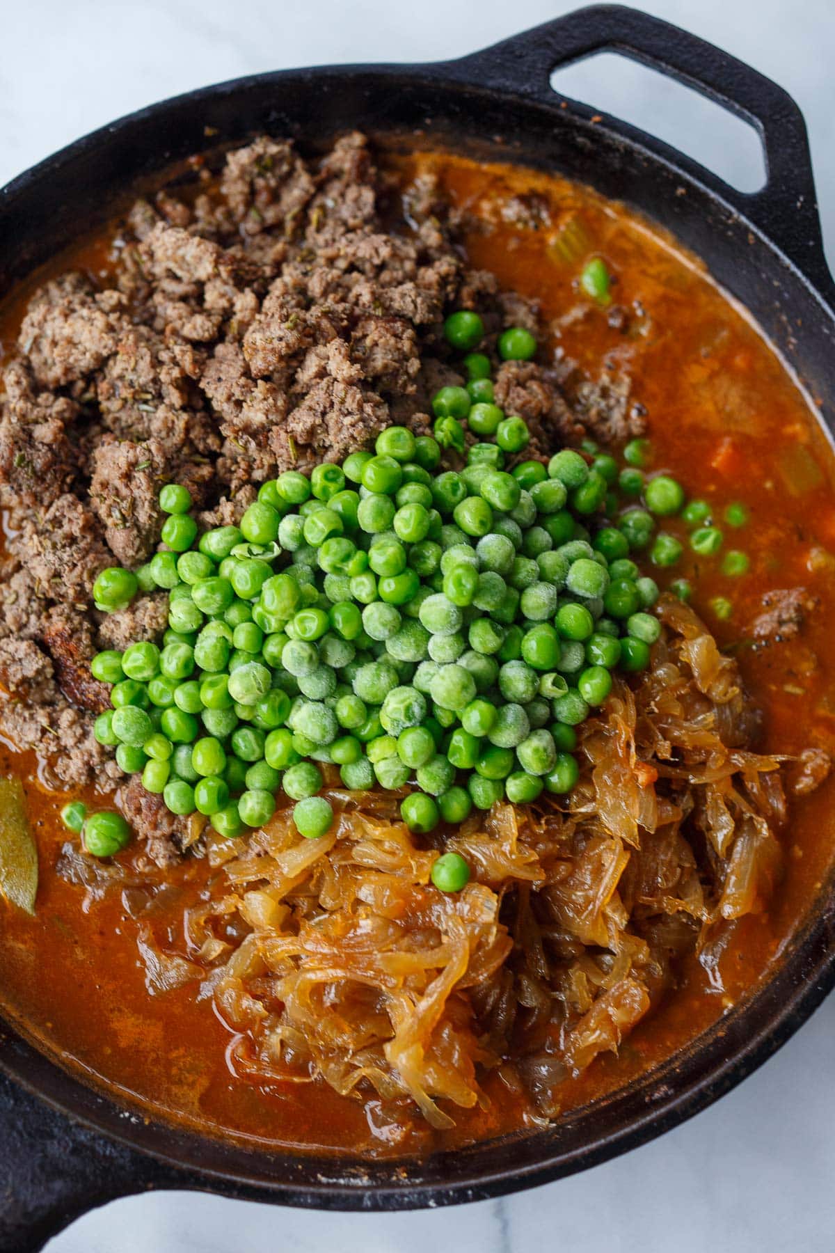 cast iron skillet with cottage pie filling coming together - ground beef, peas, caramelized onions, broth, and veggies. 