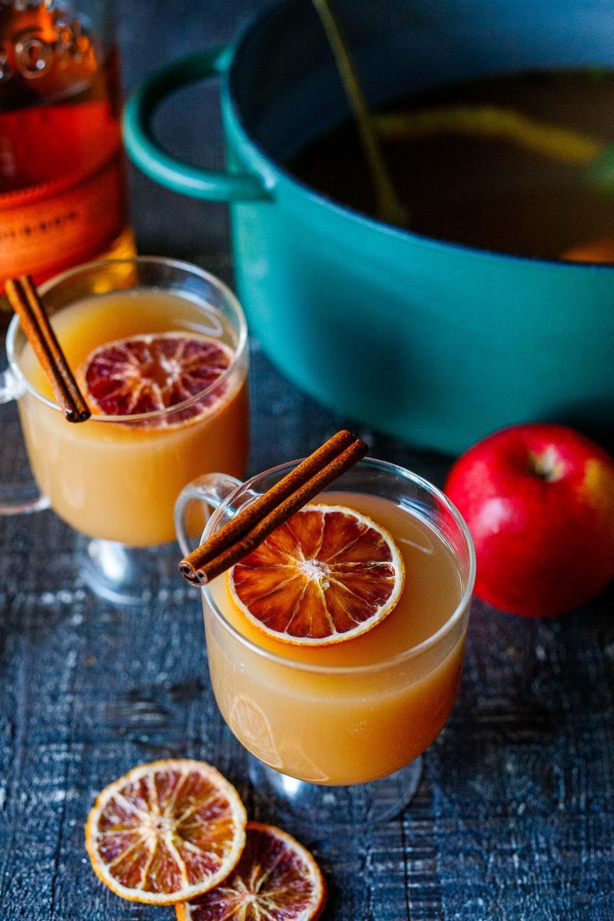 glass mugs with hot bourbon apple cider topped with dried orange slice with cinnamon stick on the rim.