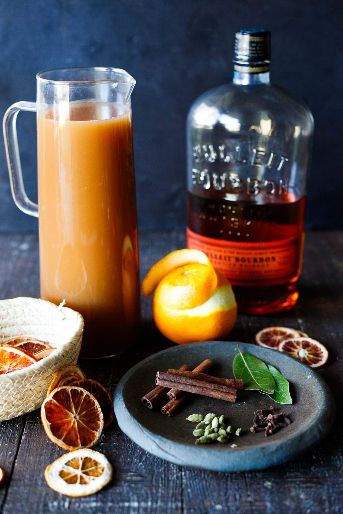 ingredients for hot bourbon apple cider - glass pitcher of apple cider, bottle of bourbon, orange with zest peeling, dish of cinnamon sticks, bay leaves, cardamom pods, and cloves, woven basket of dried orange slices.