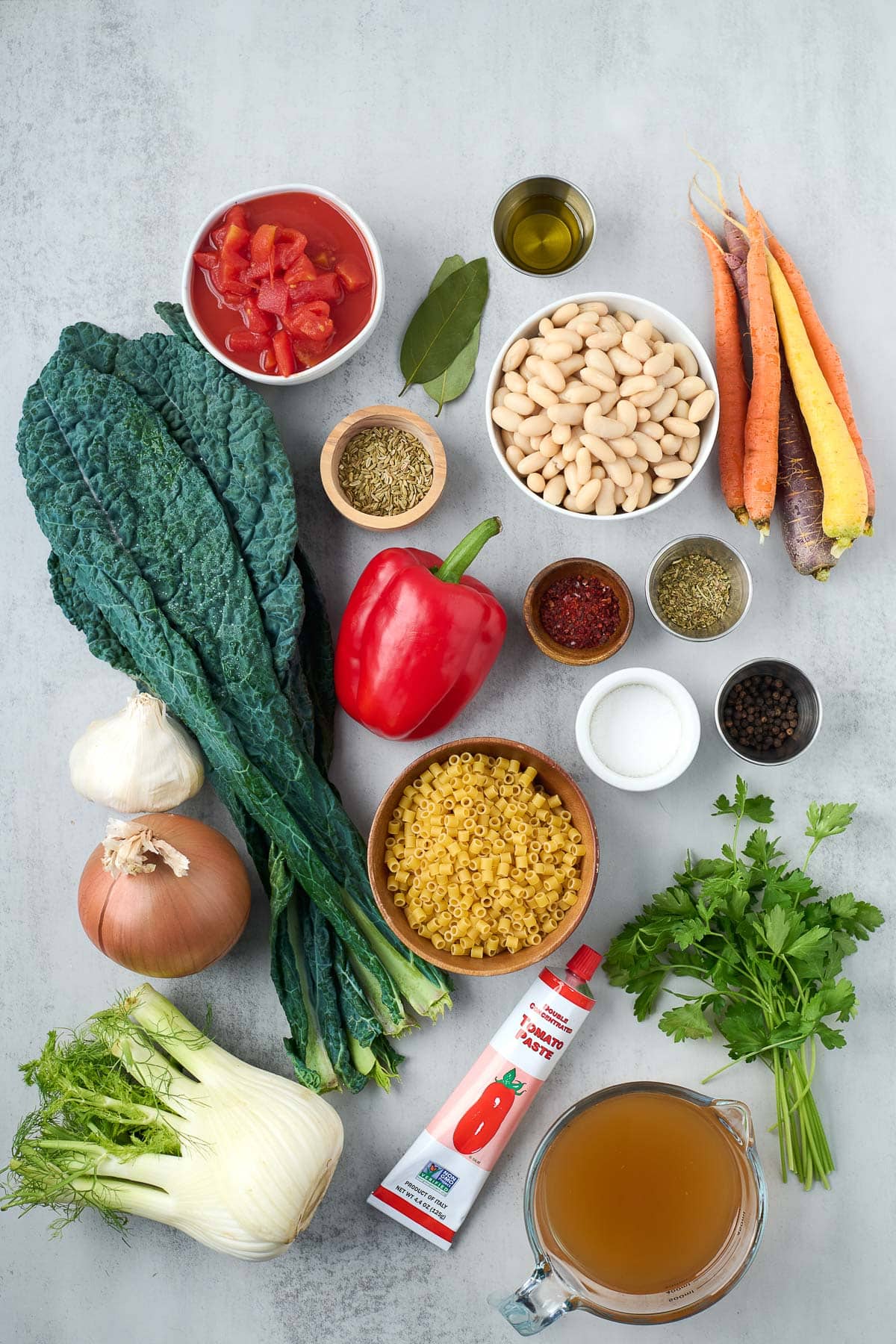 ingredients neatly arranged for minestrone soup - diced tomatoes, bay leaves, oil, fennel seeds, white beans, carrots, kale, red bell pepper, chili flakes, Italian seasoning, salt and pepper, ditalini pasta, garlic, onion, fennel bulb, tomato paste, parsley, and vegetable broth.