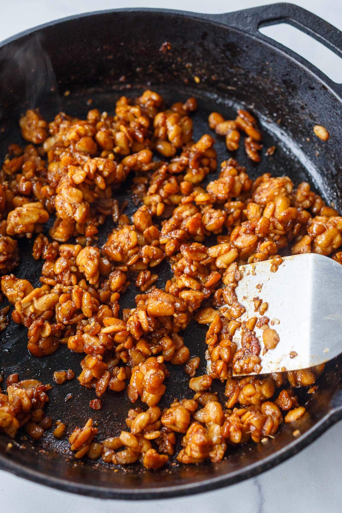 cast iron skillet with metal spatula stirring sticky tempeh. 