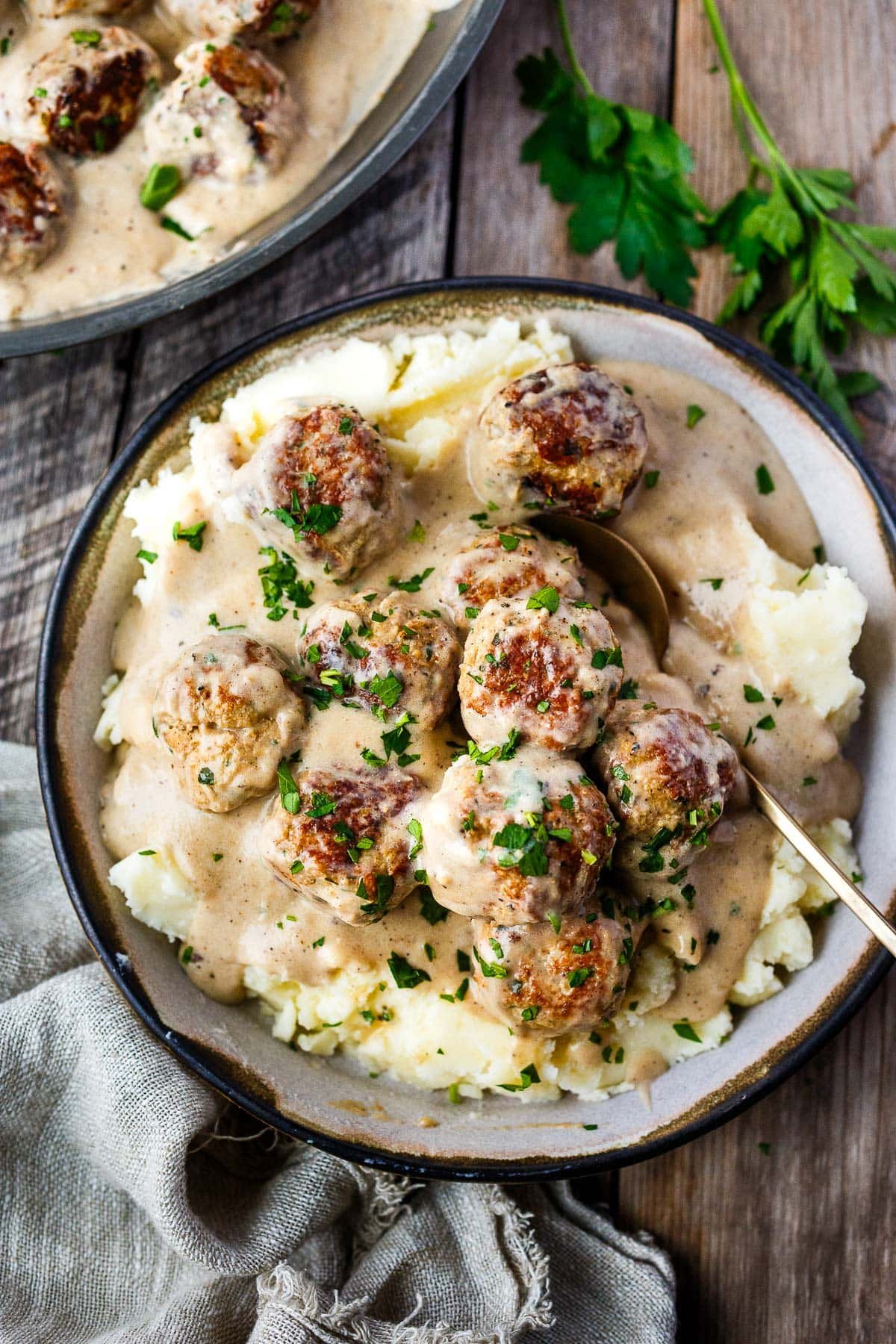 serving bowl with Swedish meatballs in gravy over mashed potatoes with gold spoon and freshly minced parsley.