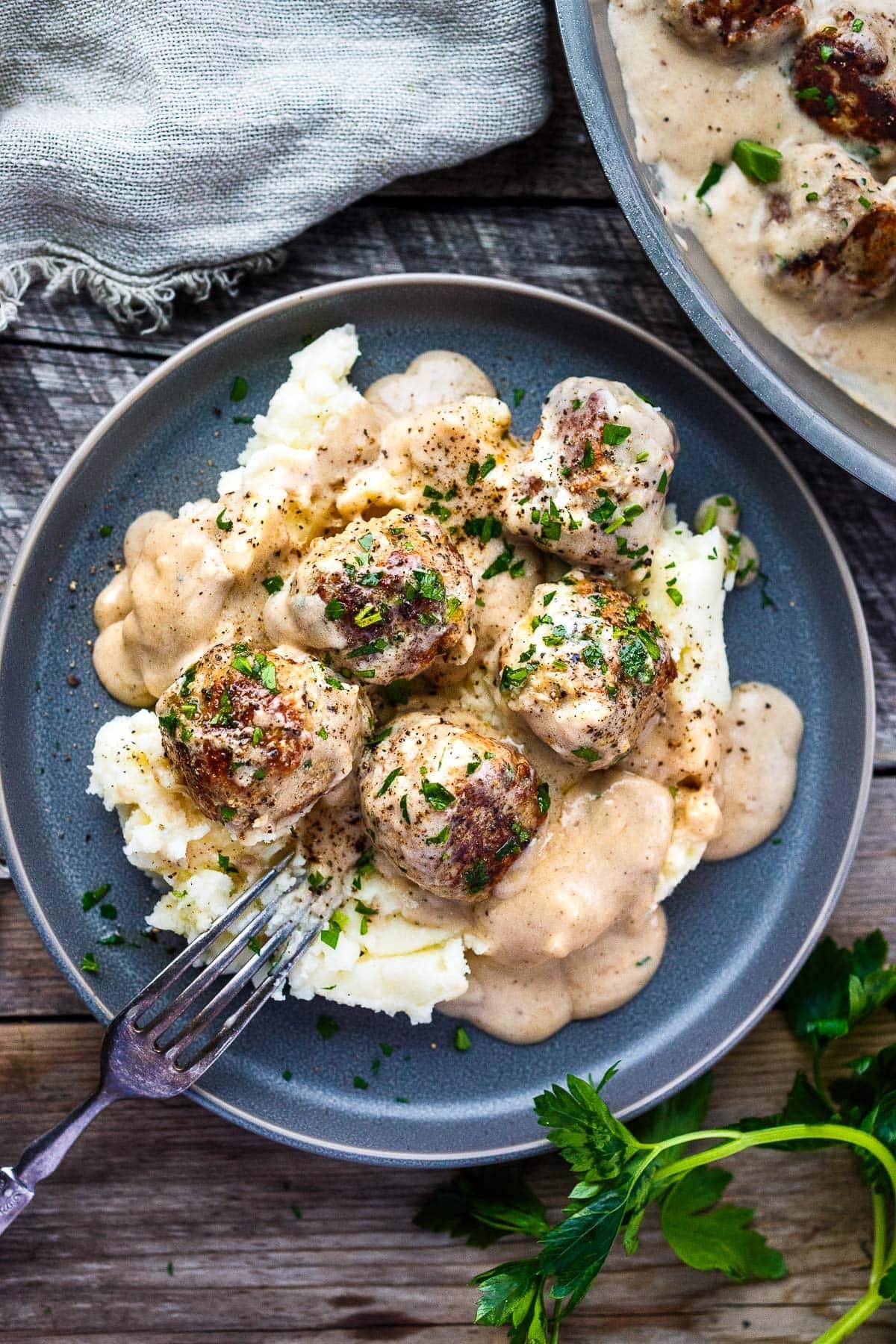 plate of swedish meatballs in gravy over mashed potatoes garnished with parsley with fork leaning upside down against plate.
