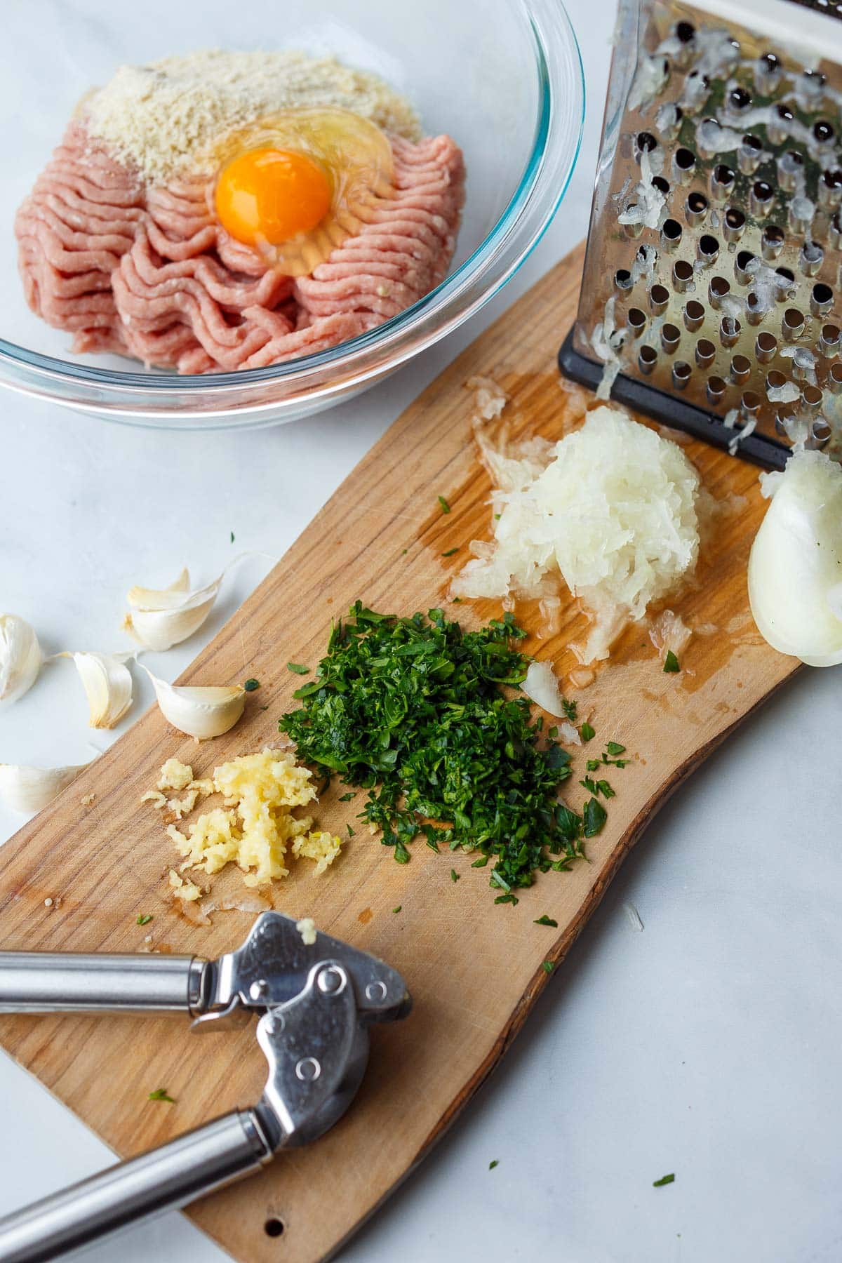 prepping Swedish meatballs with garlic press on wood board, minced parsley, and grated onion next to bowl of ground turkey with egg and breadcrumbs.