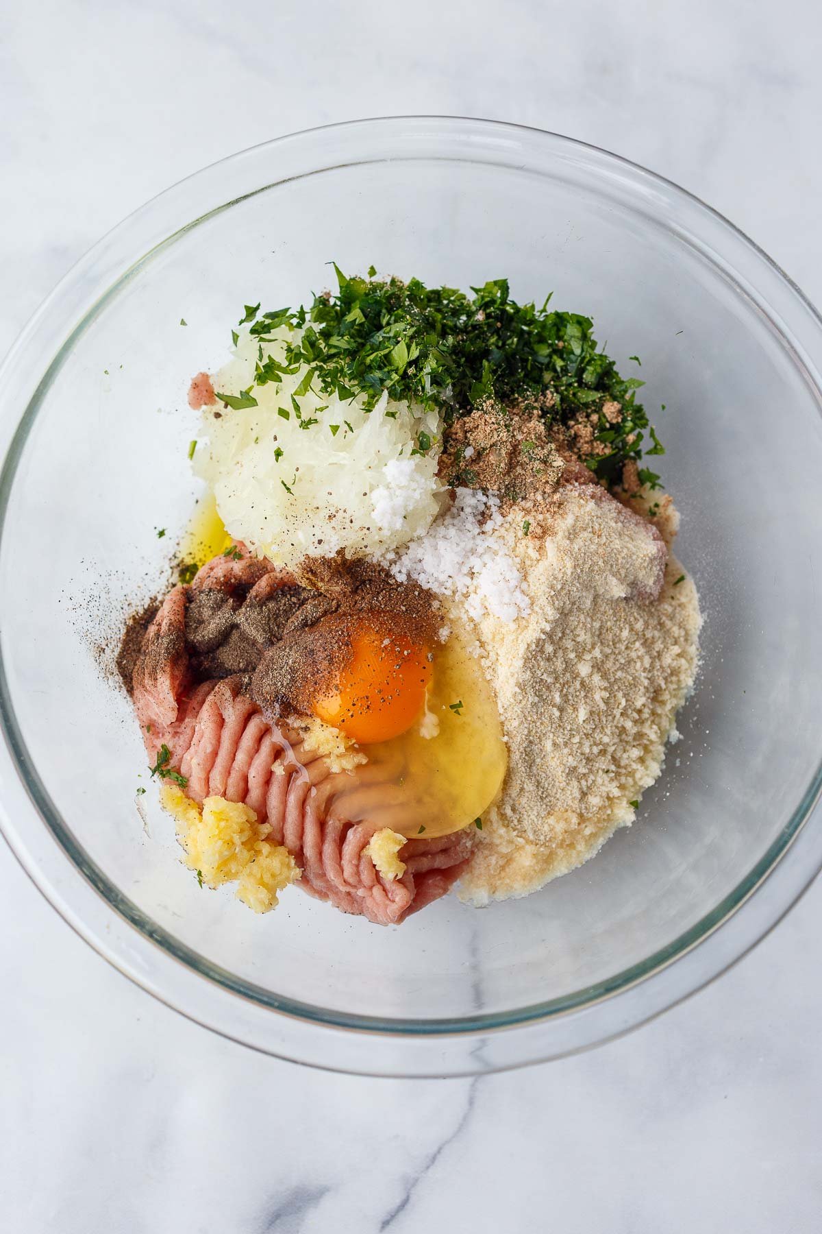 glass mixing bowl with ingredients for Swedish meatballs - egg, ground turkey, breadcrumbs, grated onion, pressed garlic, spices, and minced parsley.